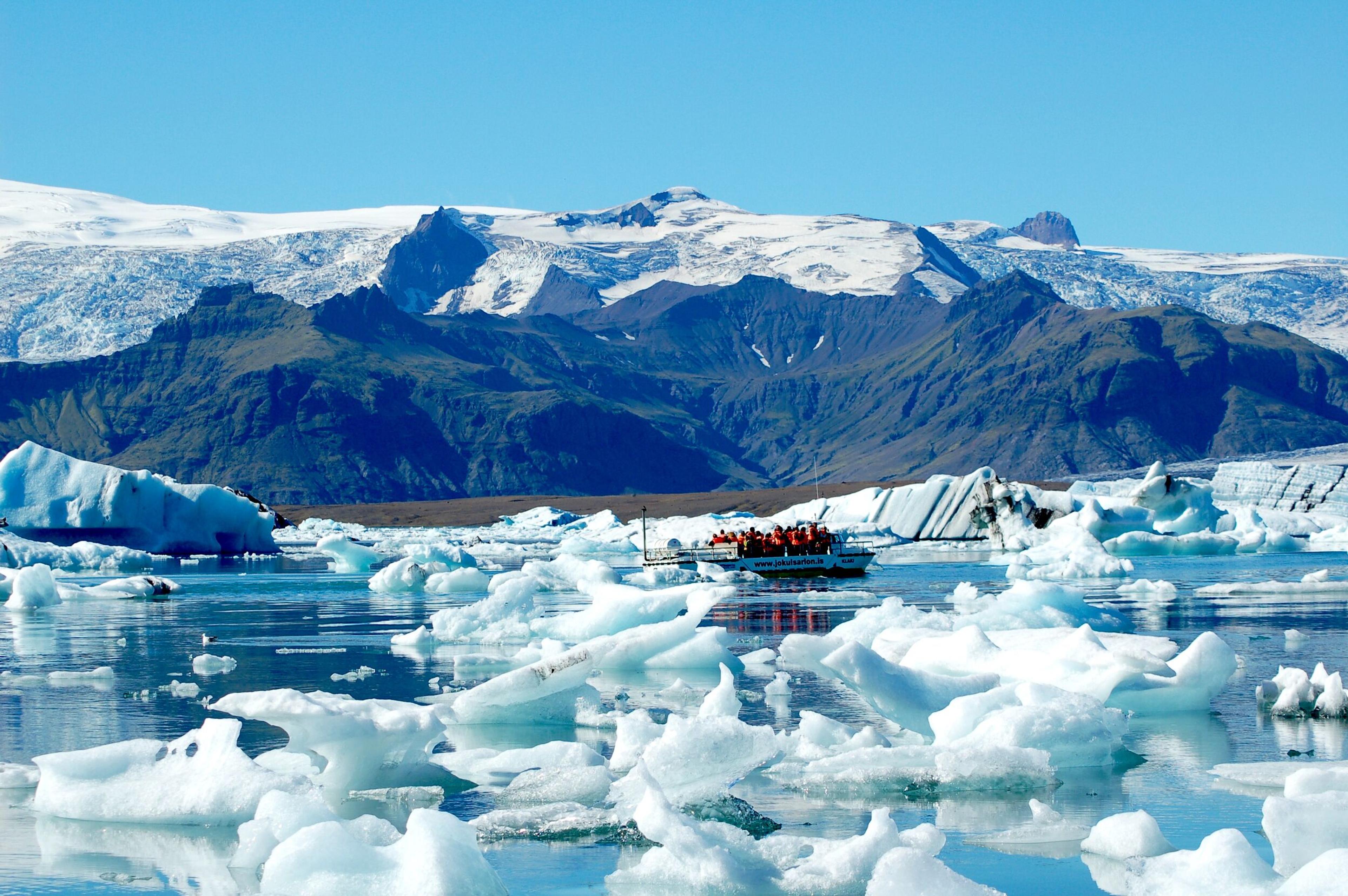 An amphibious boat floating among blue icebergs on a lake, with glacier-topped mountains in the background