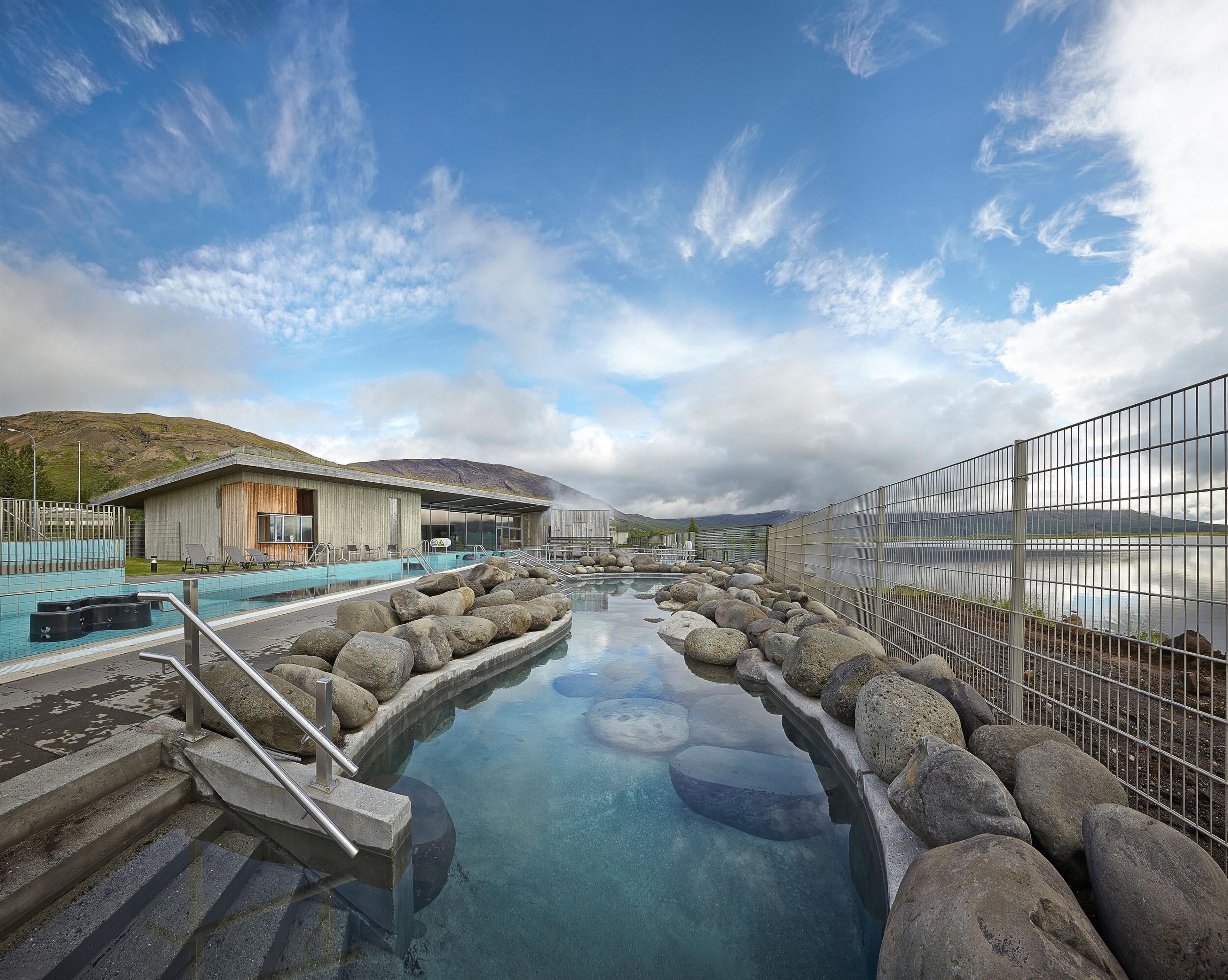 An outdoor geothermal pool with steaming blue water, nestled among rocks under a dramatic sky, featuring a wooden building in the background, symbolizing relaxation and natural beauty.