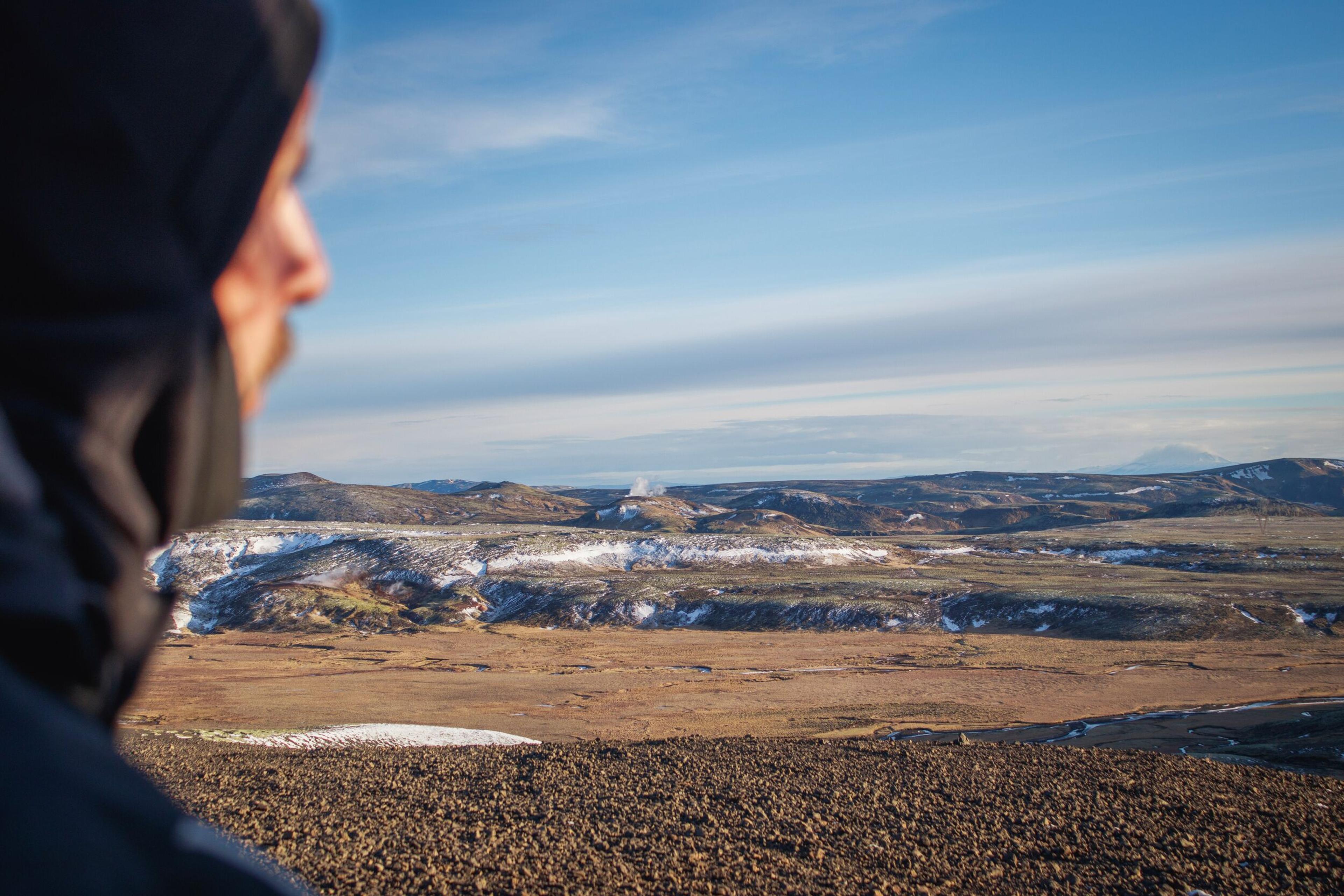 Profile of a person looking out at a vast, partially snow-covered volcanic landscape with steam rising from a distant hill.