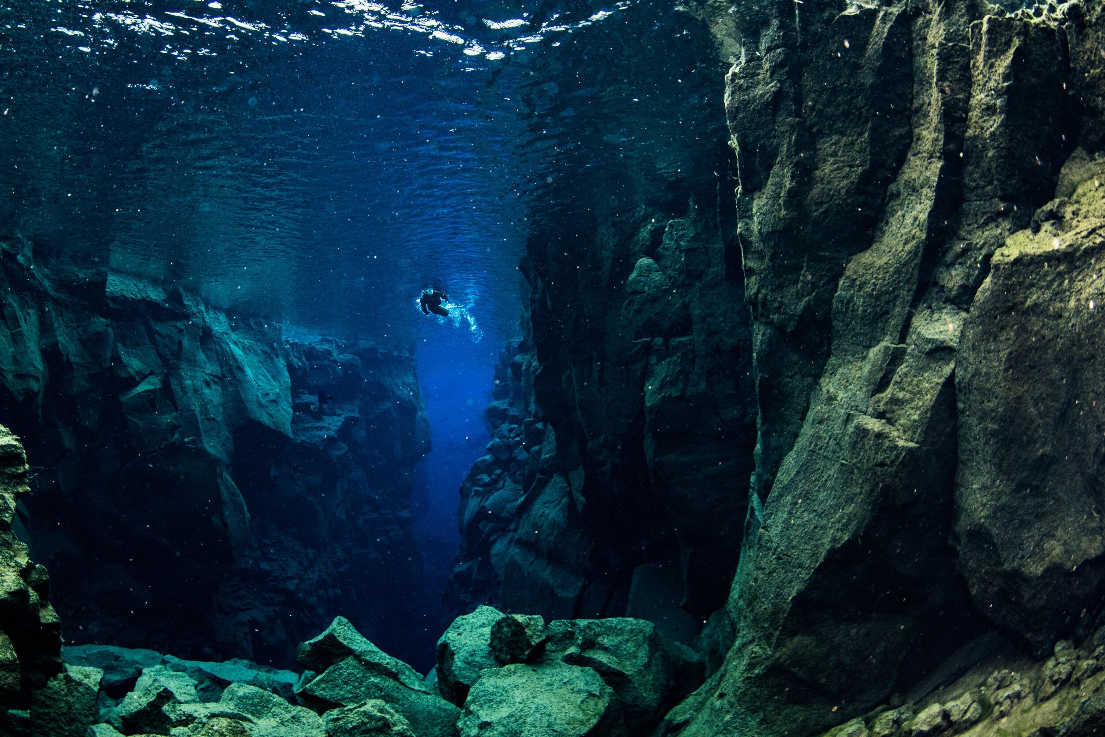 An underwater view of a diver exploring the clear, deep blue waters of a fissure between dark, jagged rock faces, creating an ethereal underwater landscape.