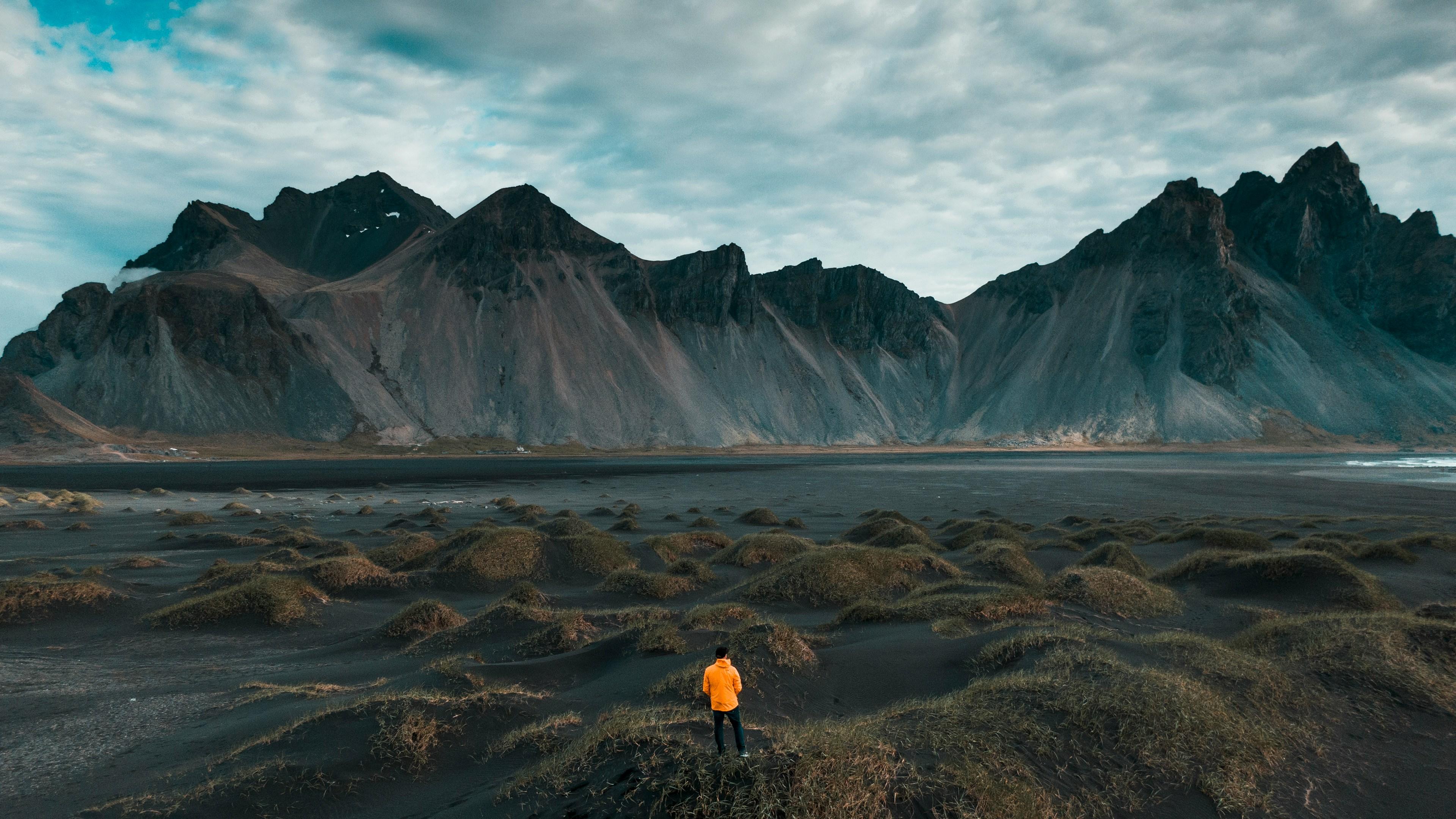 A person in an orange jacket stands on a black sand landscape with grassy mounds, facing towering dark mountains under a cloudy sky.
