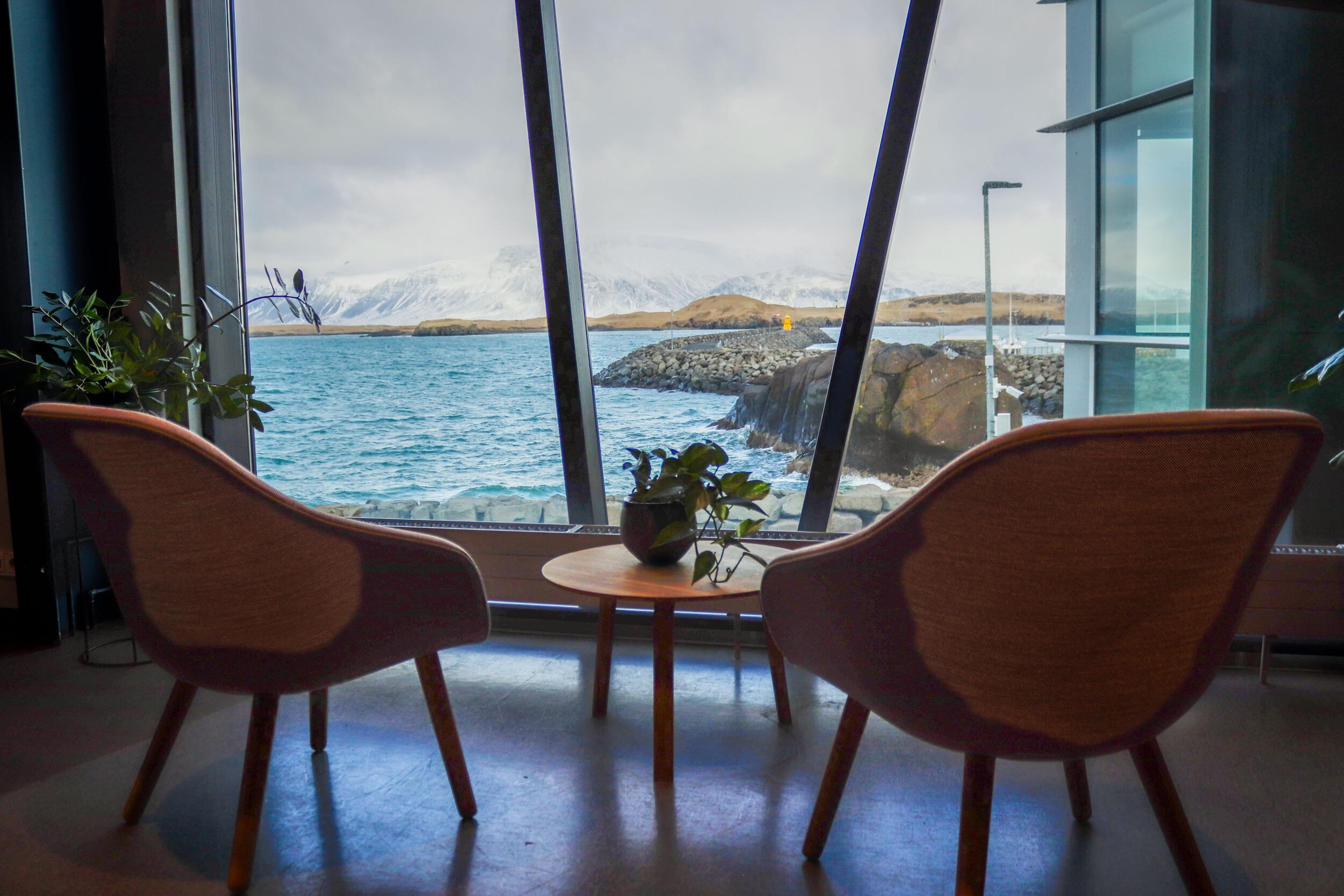 Beautiful view through a window with a focus on a table set with two chairs in the room, a small plant in the center, and a pair of glasses, suggesting a cozy spot for a meeting or relaxation with the same picturesque view of the sea and mountains.