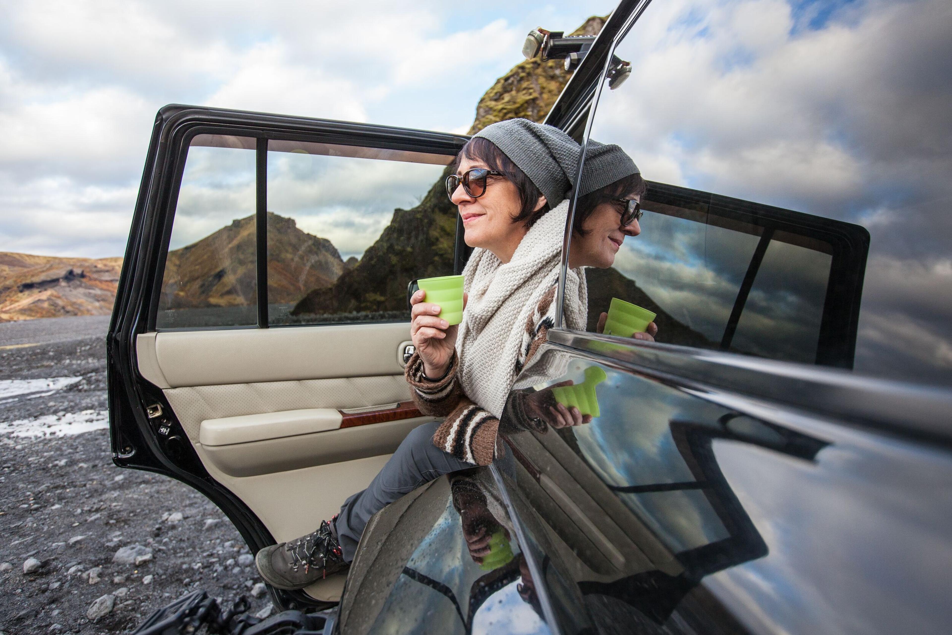 Traveler in sunglasses holding a cup, seated inside a car with the door open, gazing outwards.