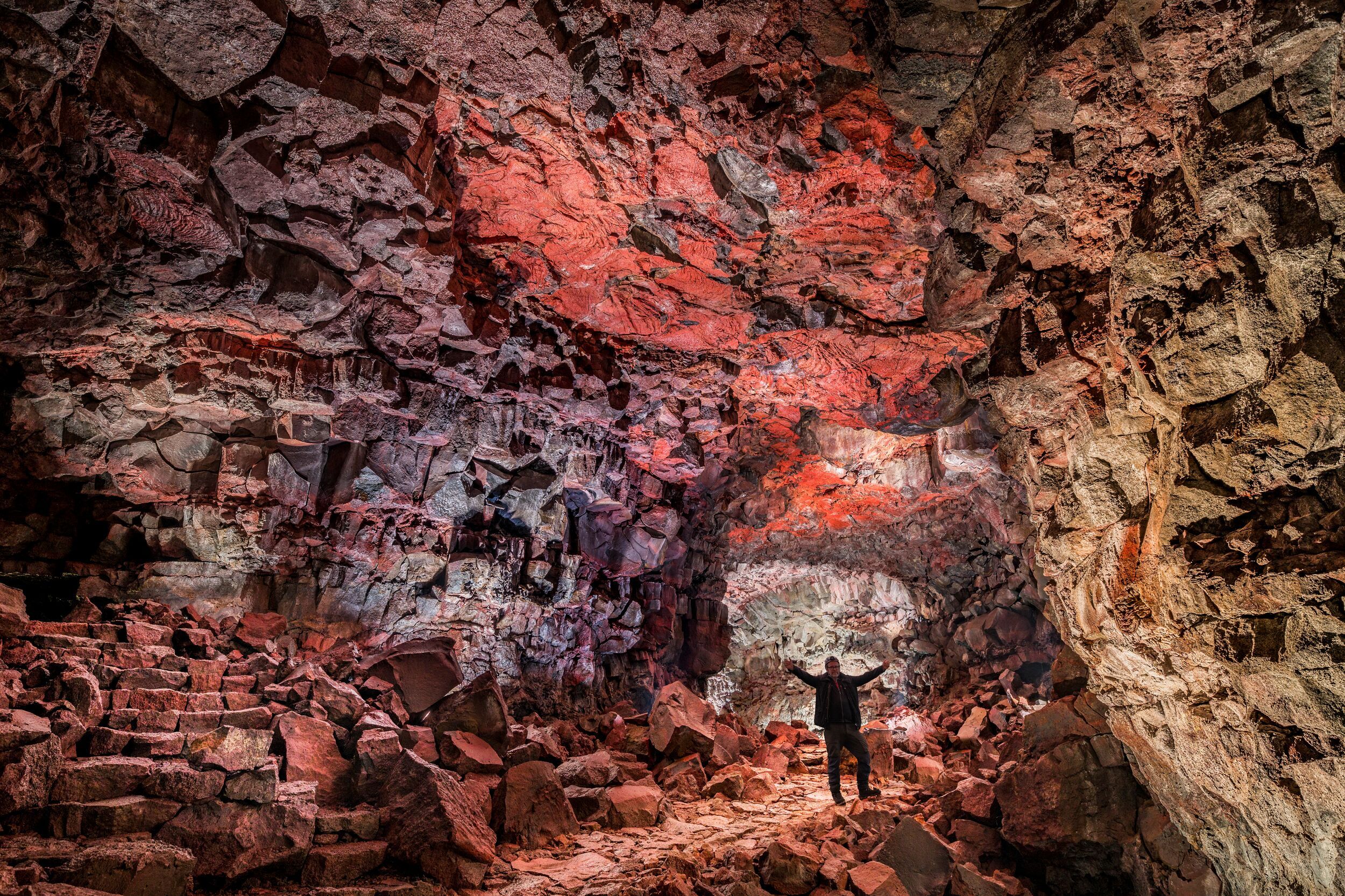 A person stands with arms raised inside a vast Iceland lava cave, surrounded by jagged red-tinged rock walls illuminated dramatically by artificial light.