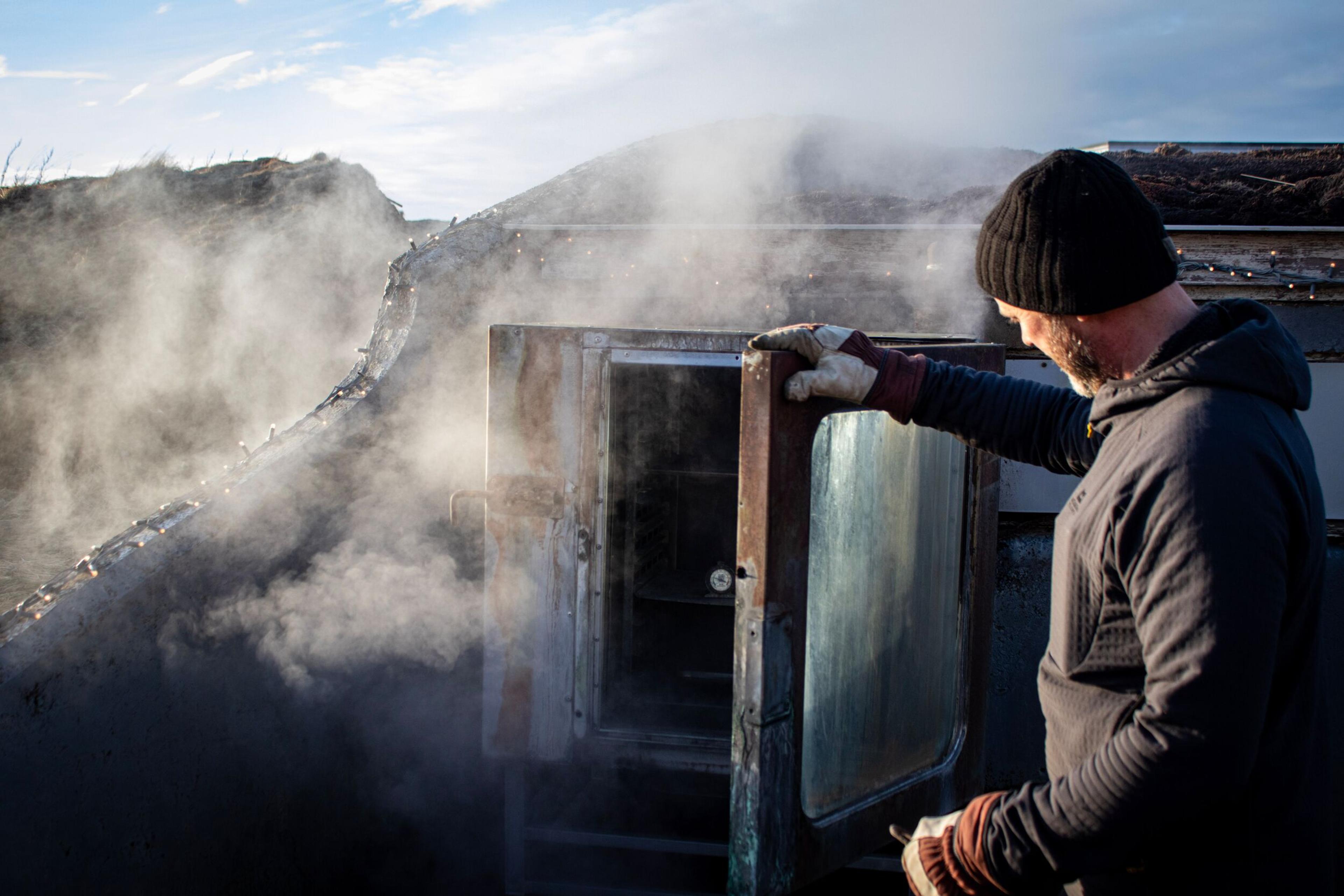 A man in a black beanie and gloves opens a steaming outdoor smoker.