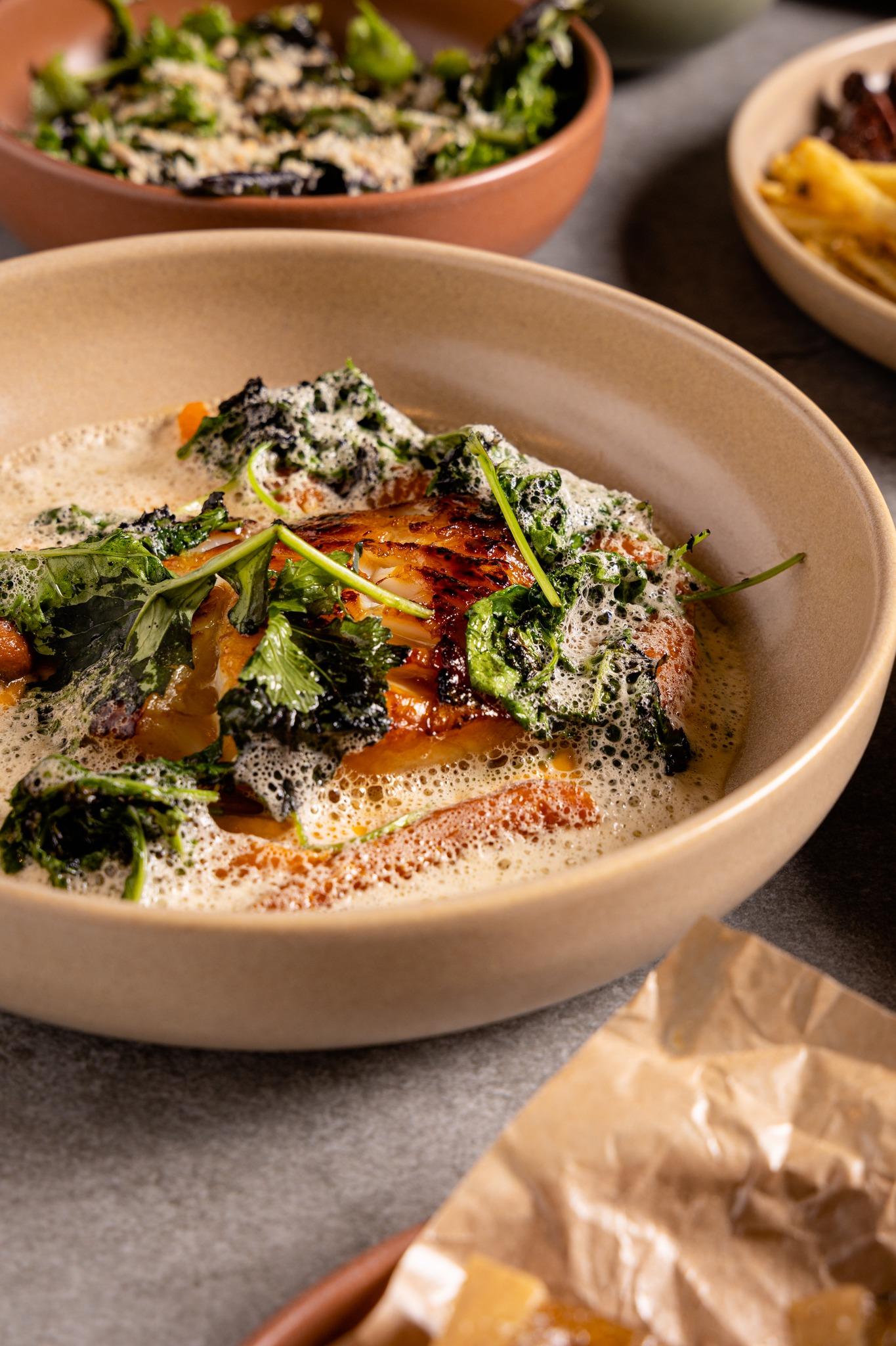 Gourmet dish at Hafnartorg Gallery Food Hall featuring seared fish with leafy greens and a creamy foam garnish in a beige bowl.