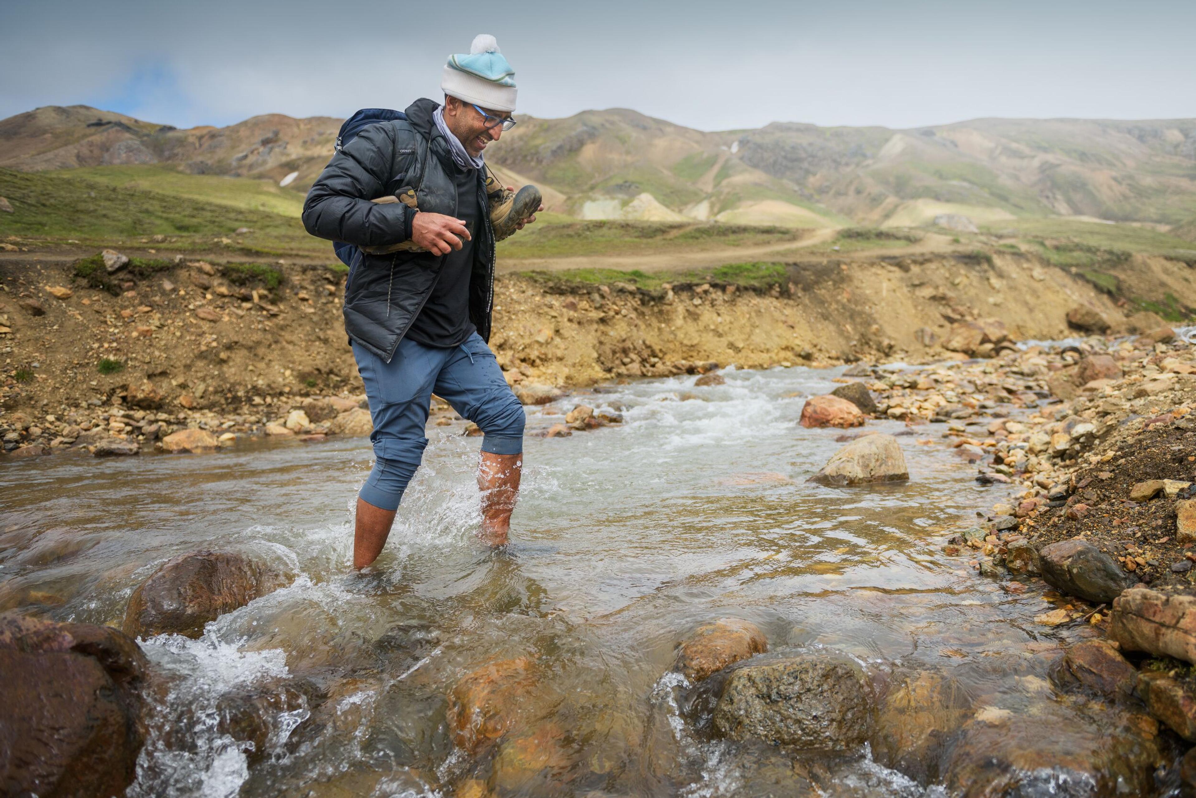 A smiling person crosses a rocky stream barefoot, holding their boots, with a mountainous landscape behind.