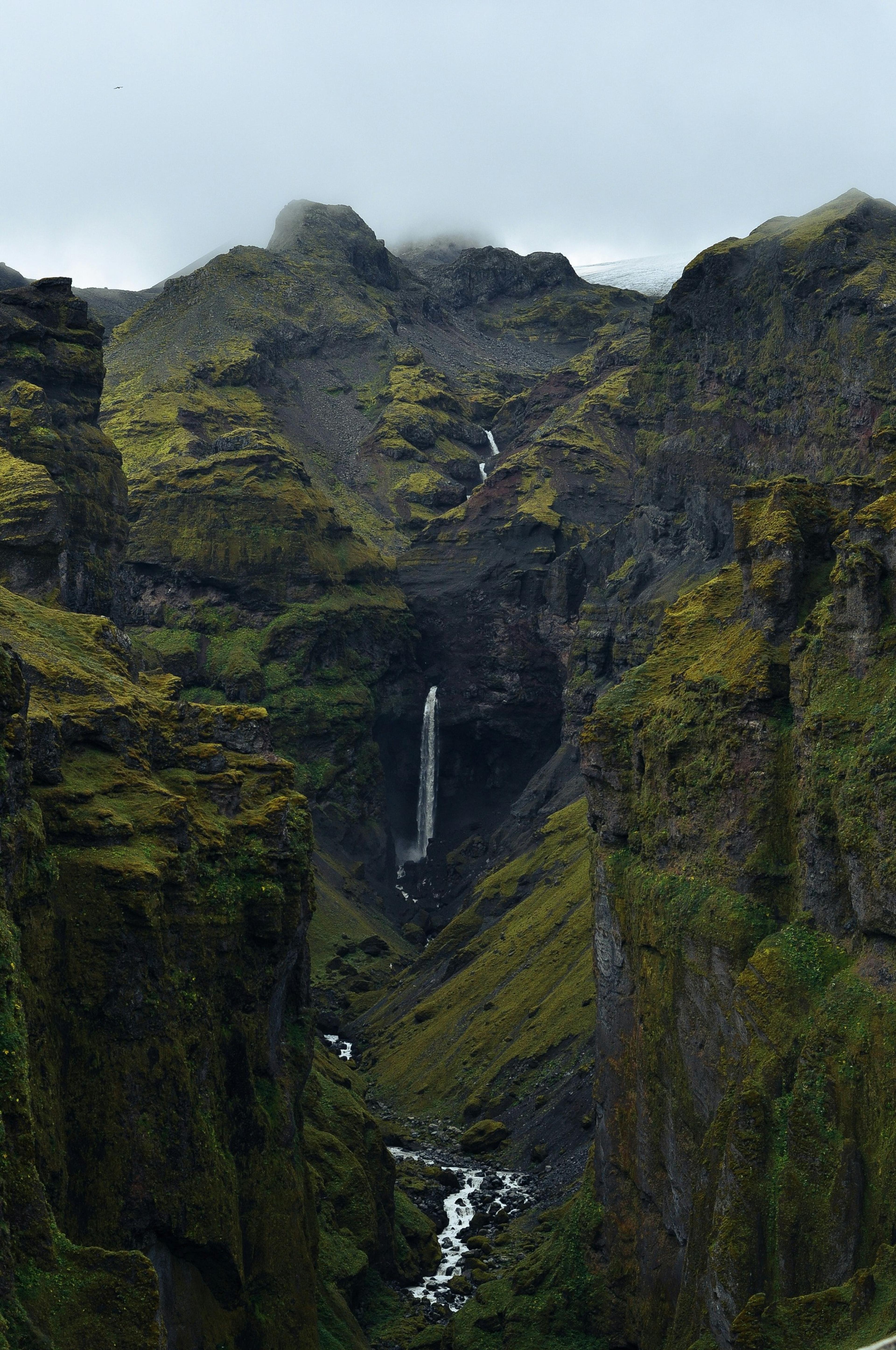 View of Mulagljufur Canyon with a vertical waterfall descending into a lush, green gorge, shrouded in mist.