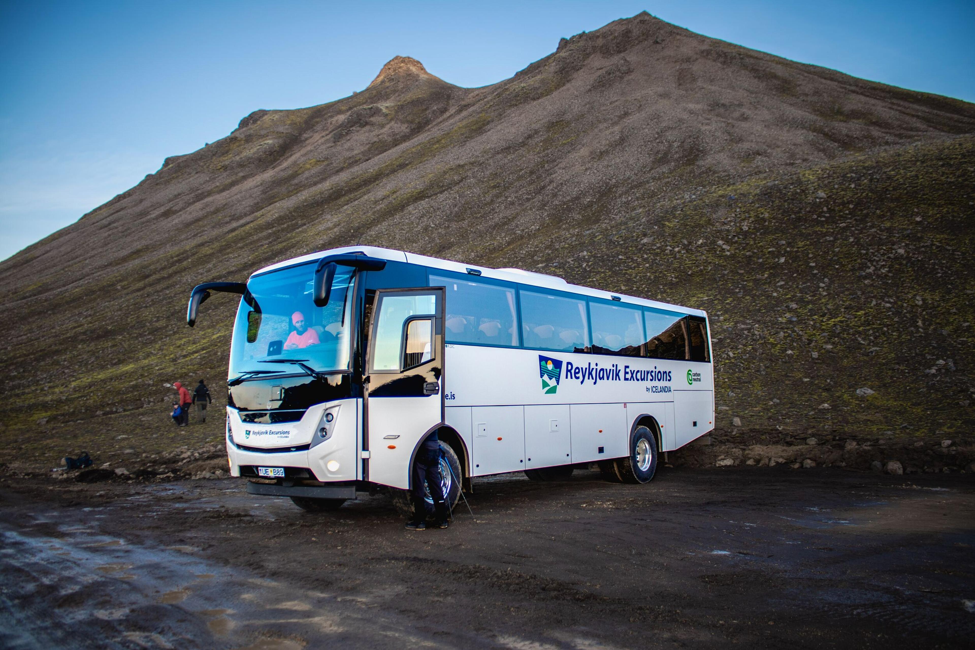 A white Reykjavik Excursions bus parked on a dirt road in front of a large, barren mountain.
