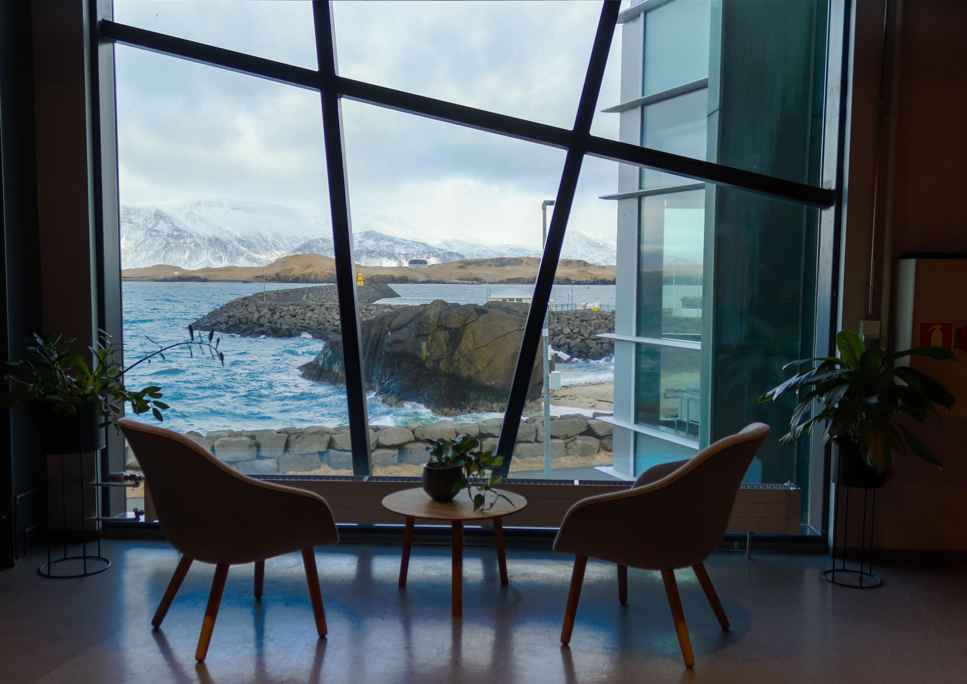  room with modern furnishings including two chairs and a table, set against floor-to-ceiling windows that showcase a rocky coastline and calm sea against a backdrop of distant mountains, under a cloudy sky.