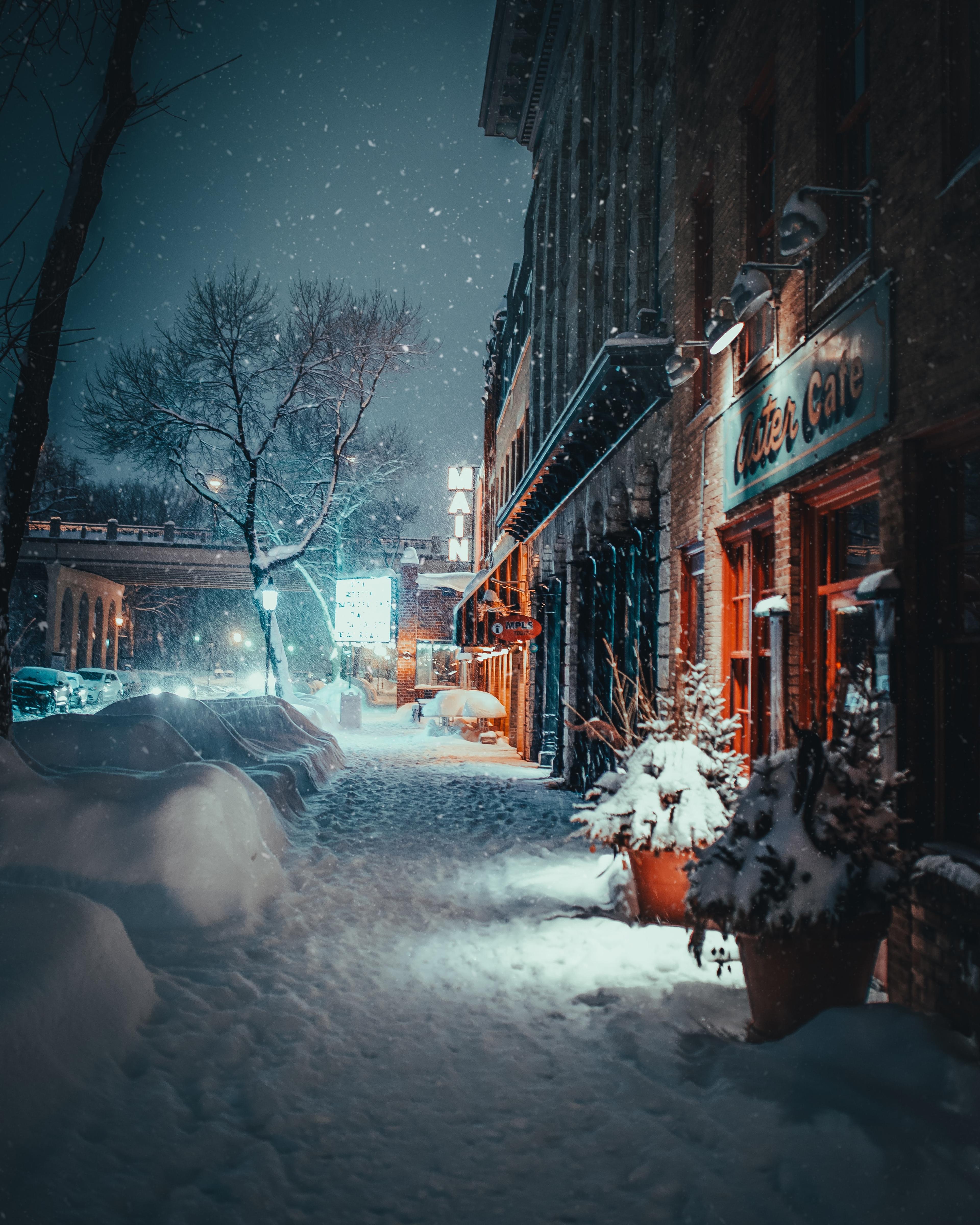 A snowy evening scene on a city street with warmly lit shop fronts, including a "Café," under a gentle snowfall, with a tranquil, holiday ambiance.