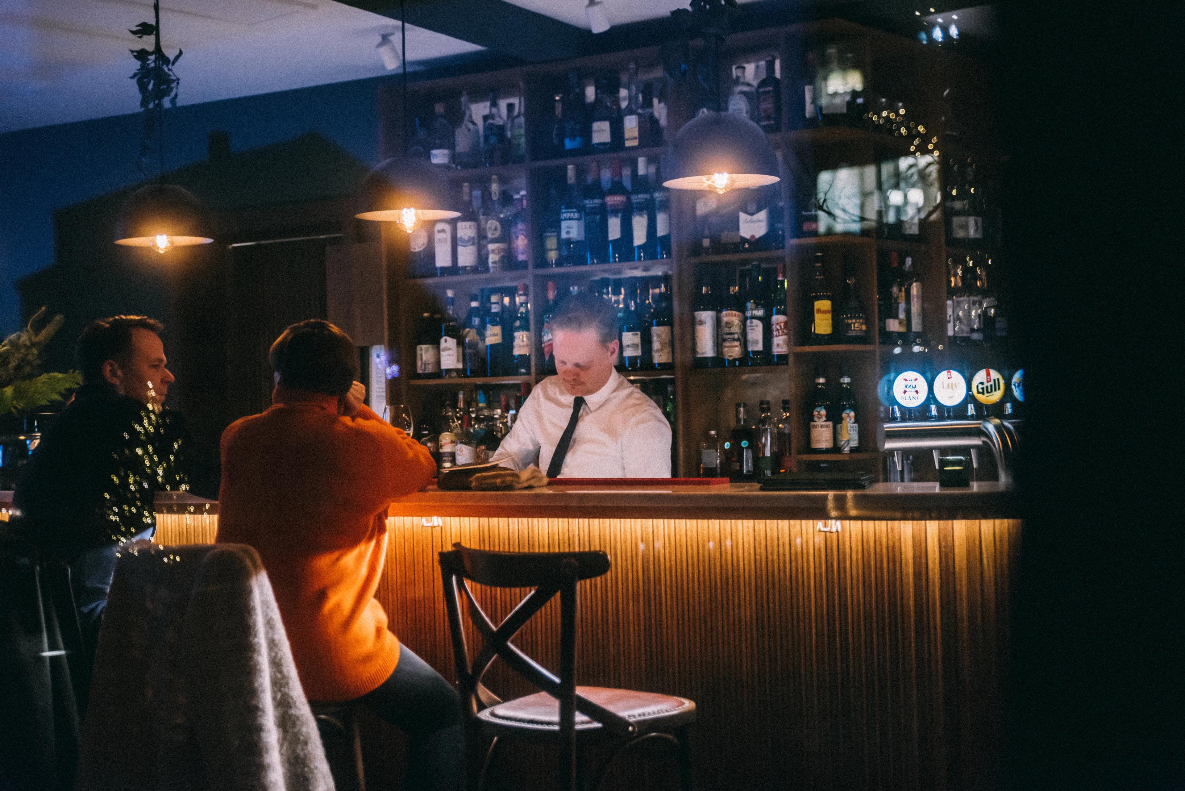 A bartender works behind a warmly lit bar as two customers sit at the counter.