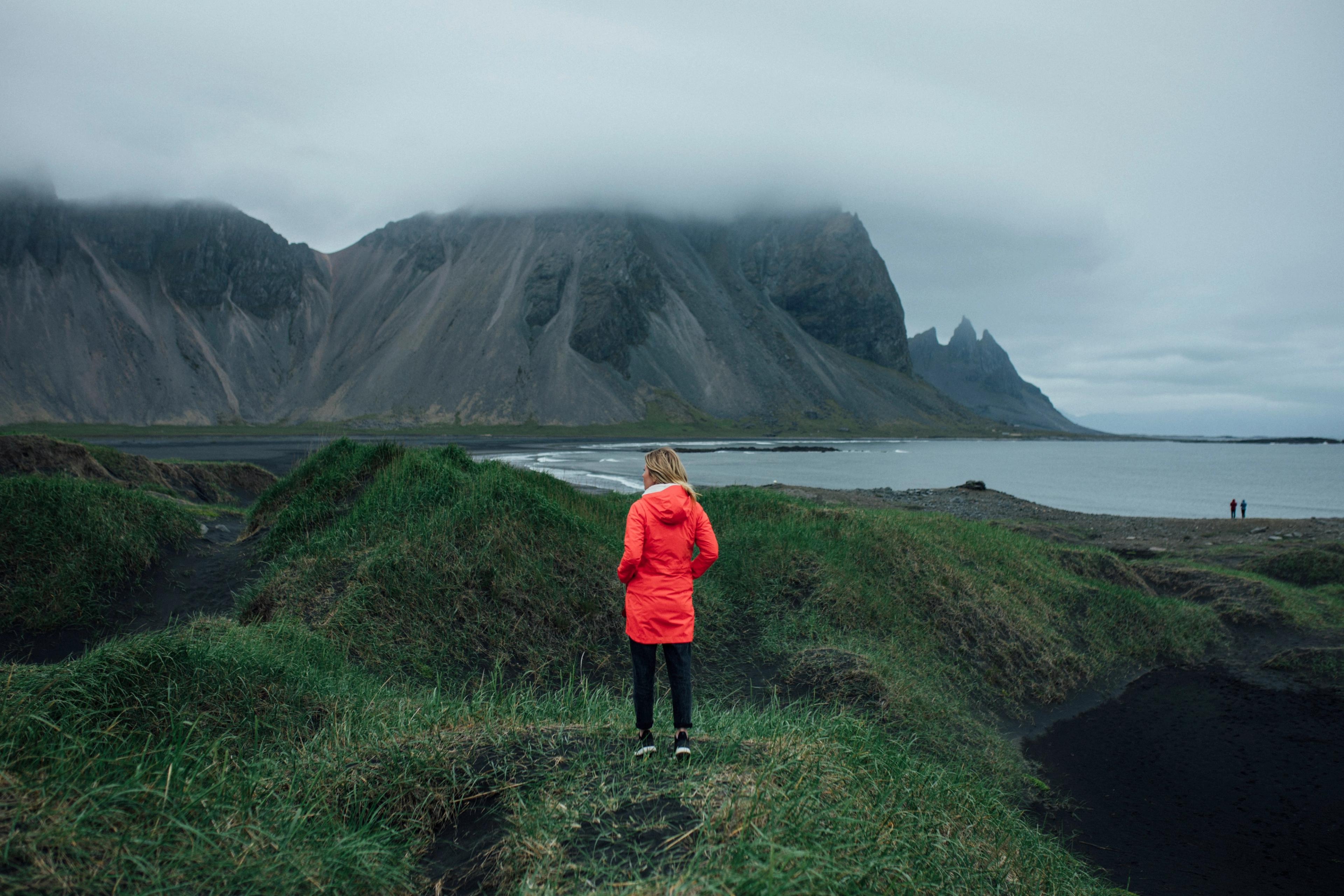 Person in a red jacket stands on a grassy hill, facing a black sand beach, dark ocean, and cloud-covered mountains.