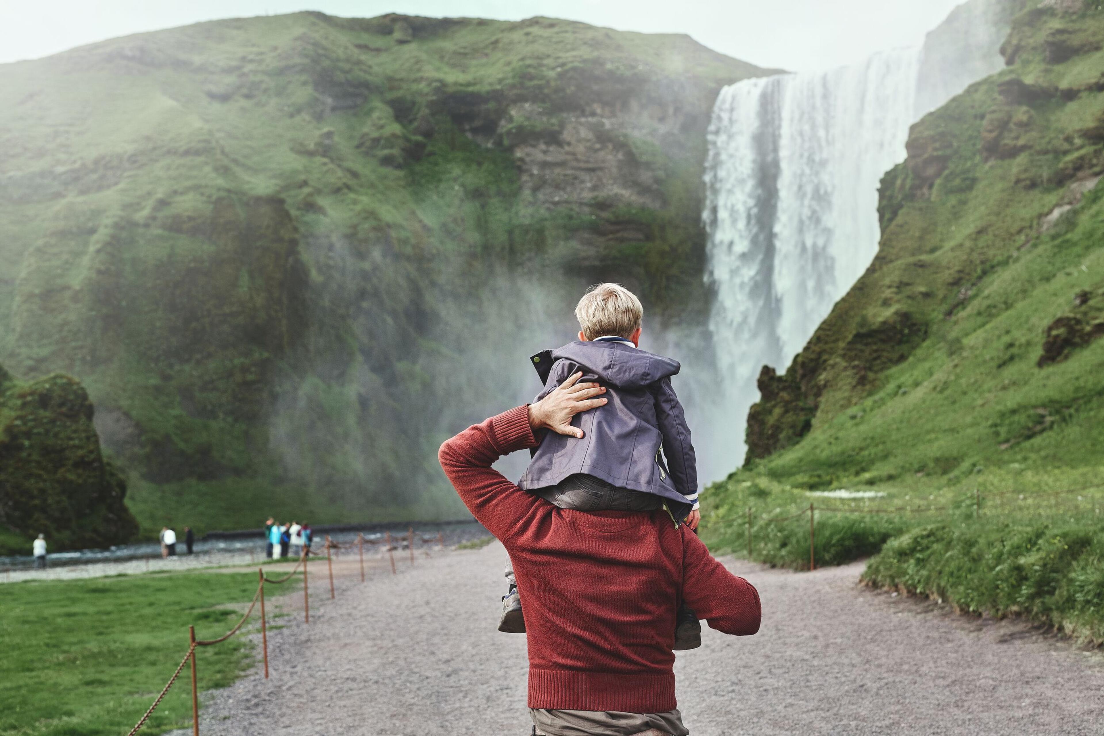 A father carrying his child on his shoulders while walking toward a large waterfall in Iceland.