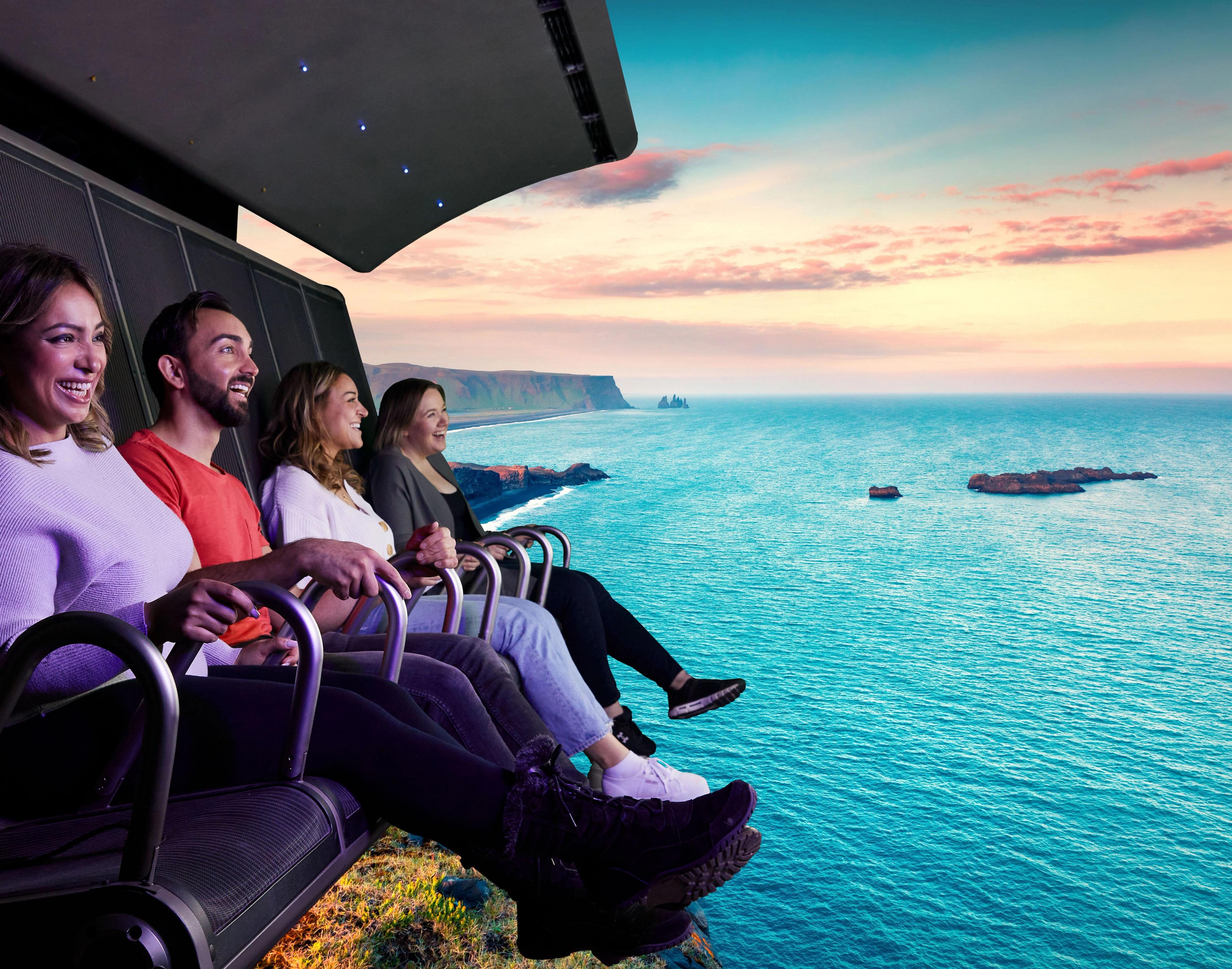 Four people on an immersive ride smiling at a dramatic coastal landscape with a turquoise ocean, cliffs, and a colorful sky.