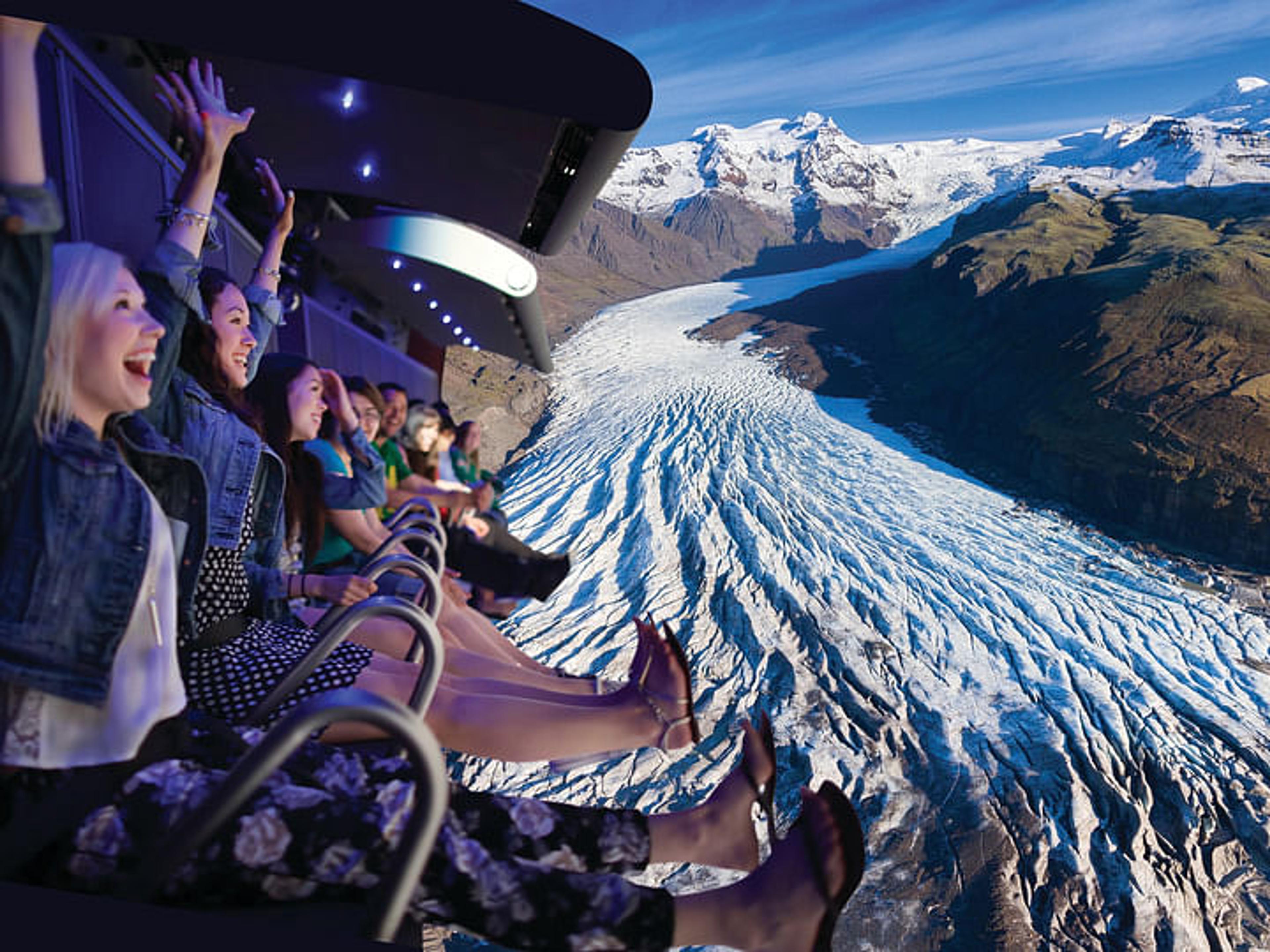 Excited people on a simulated flying ride above a glacier and mountains.