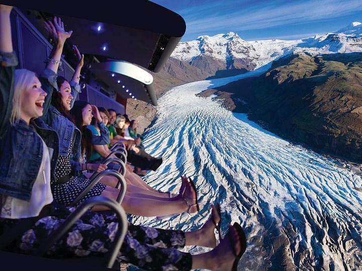 Excited people on a simulated flying ride above a glacier and mountains.