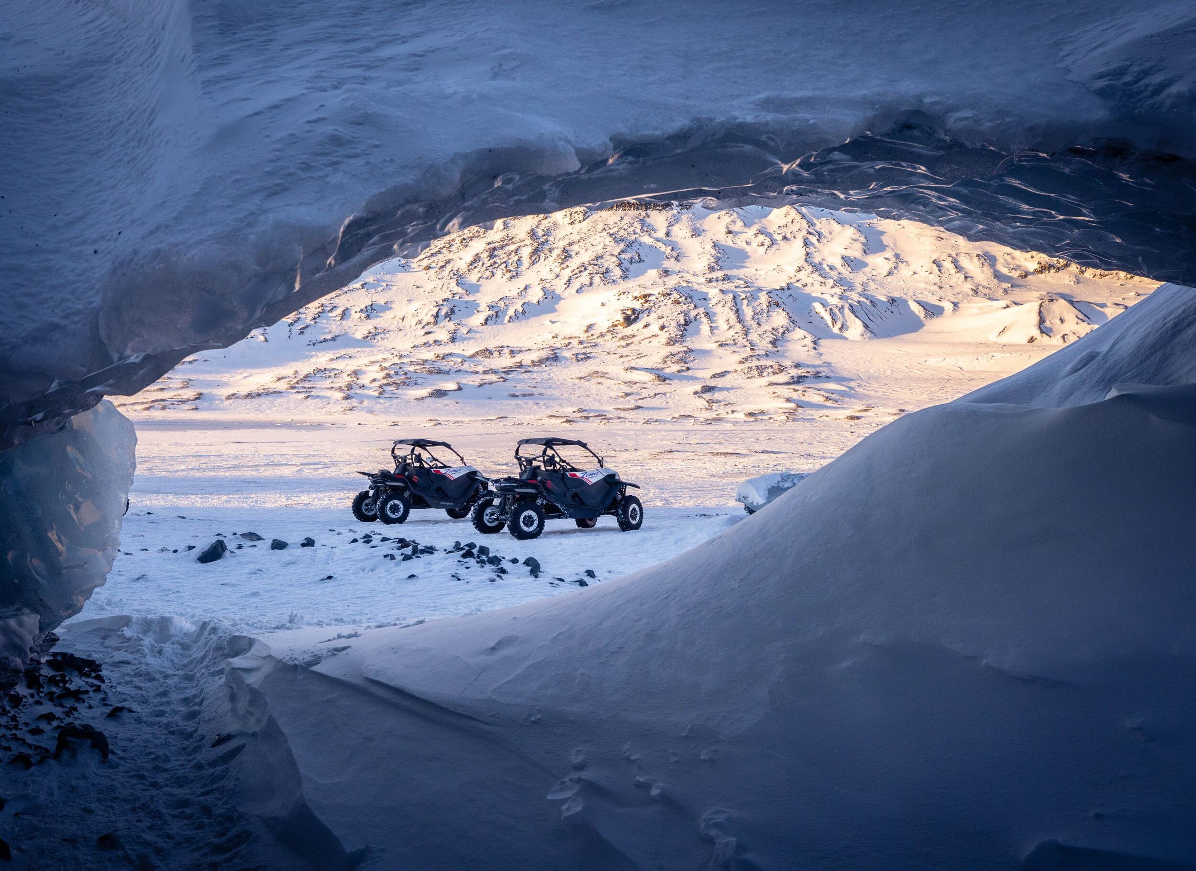 Two buggies are parked on a snowy terrain with mountains in the background, illuminated by the setting sun.