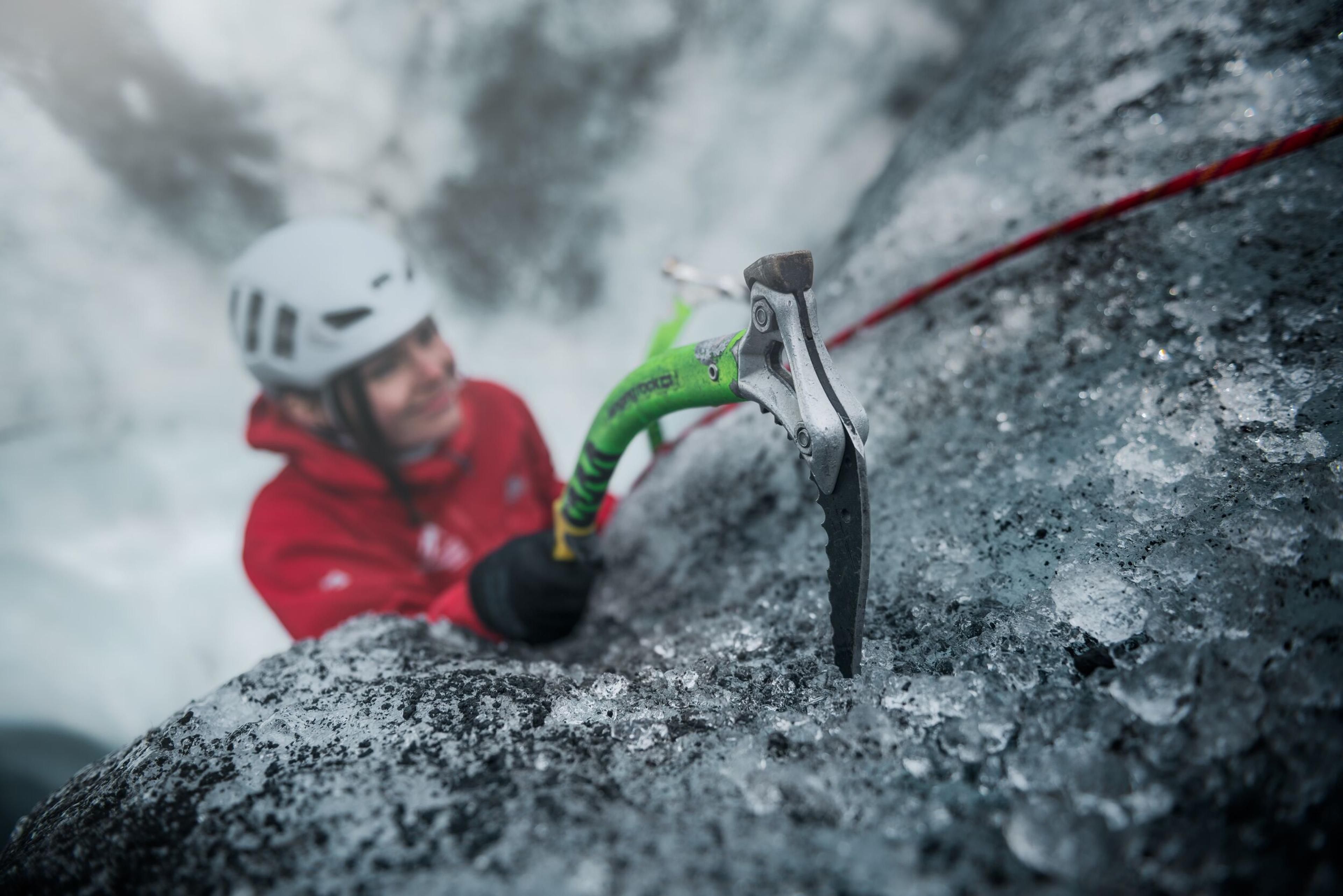 A person in a red jacket and white helmet uses an ice axe for climbing on a frost-covered rock face.