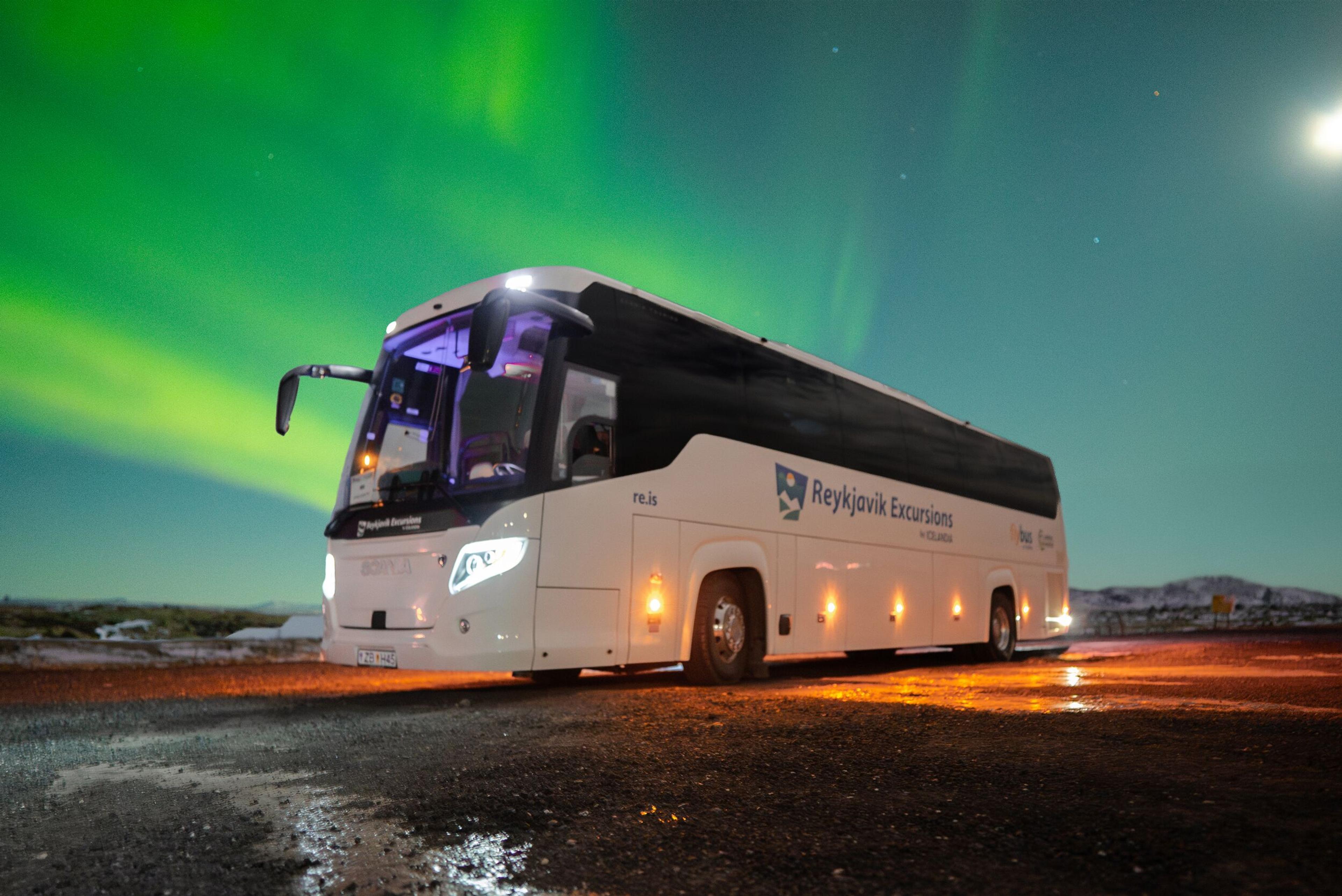 A white "Reykjavik Excursions" bus parked under the vibrant green Northern Lights at night.