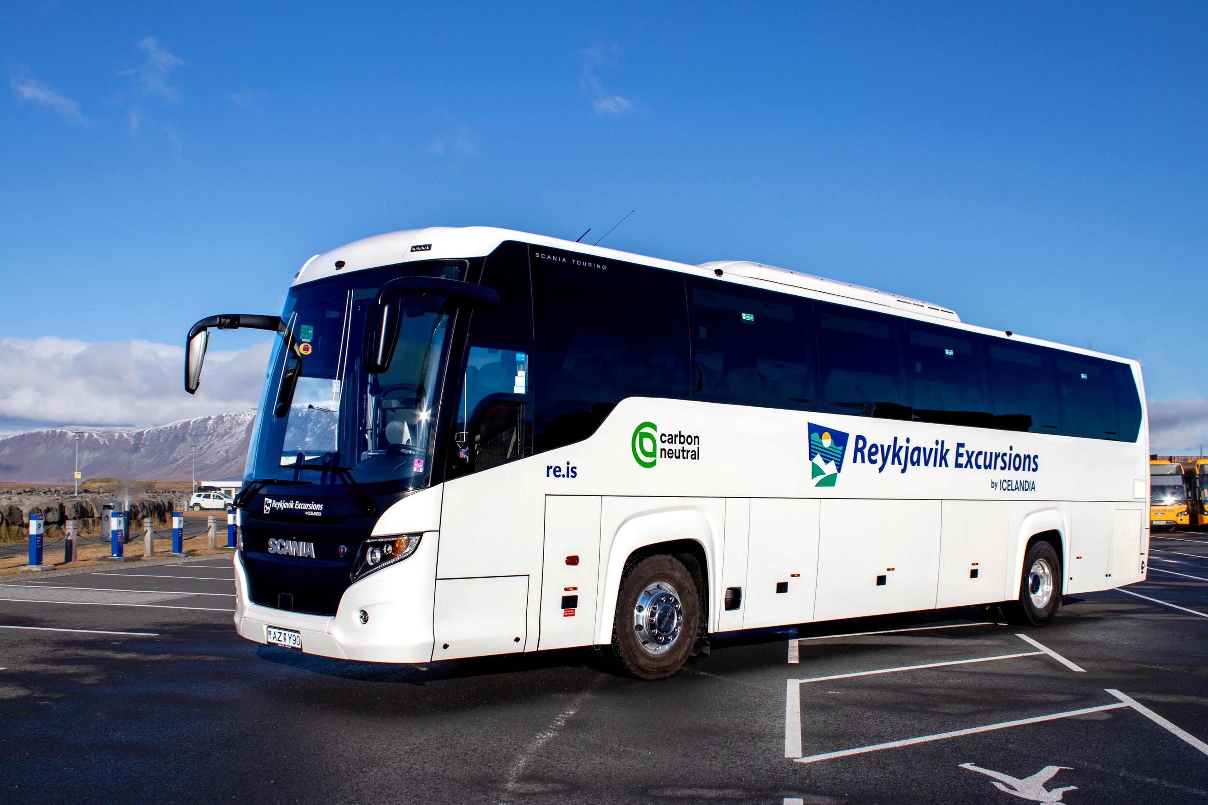 A white Reykjavik Excursions "carbon neutral" tour bus parked outdoors with mountains in the background.