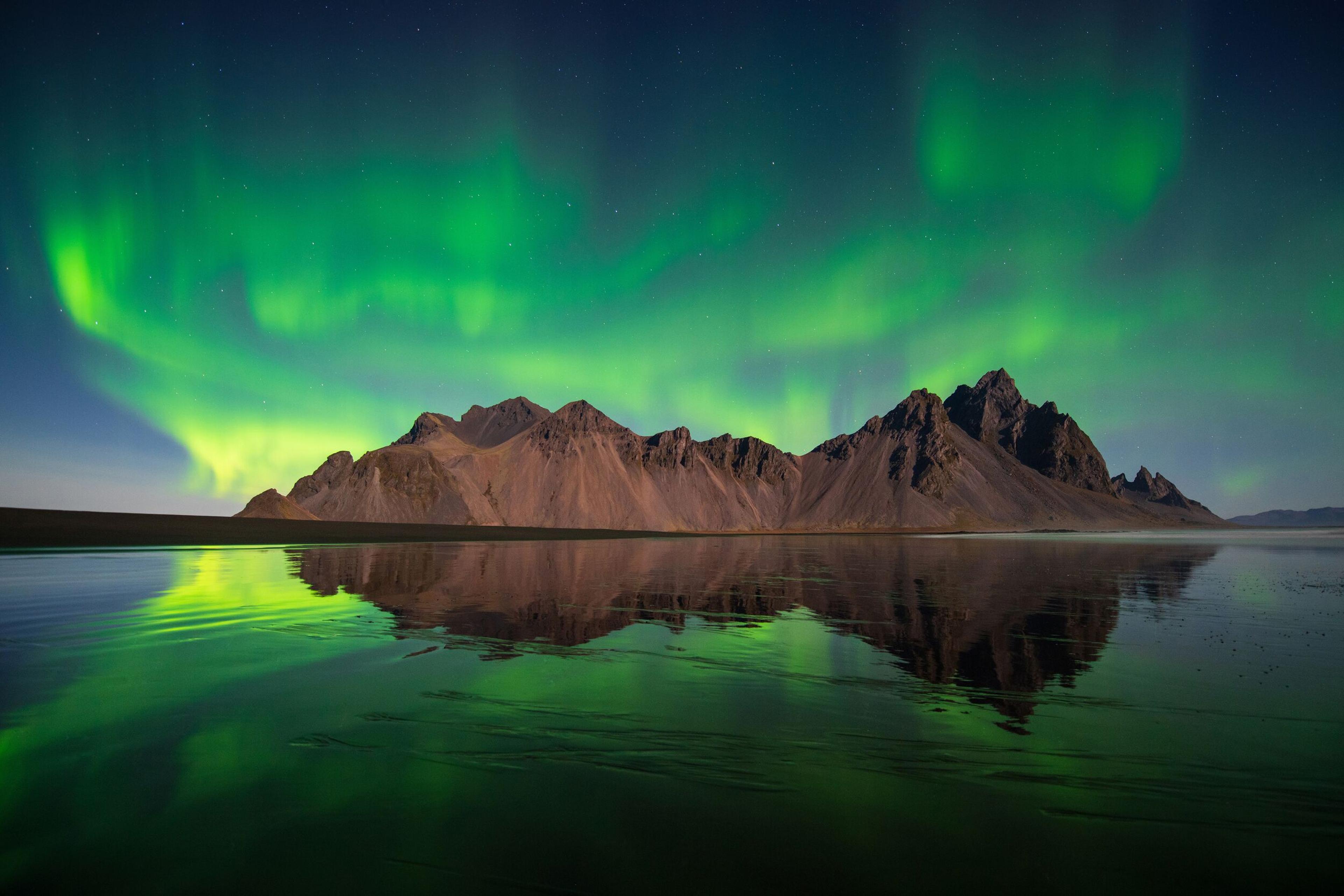 Vibrant green aurora borealis over mountains reflected in calm water.