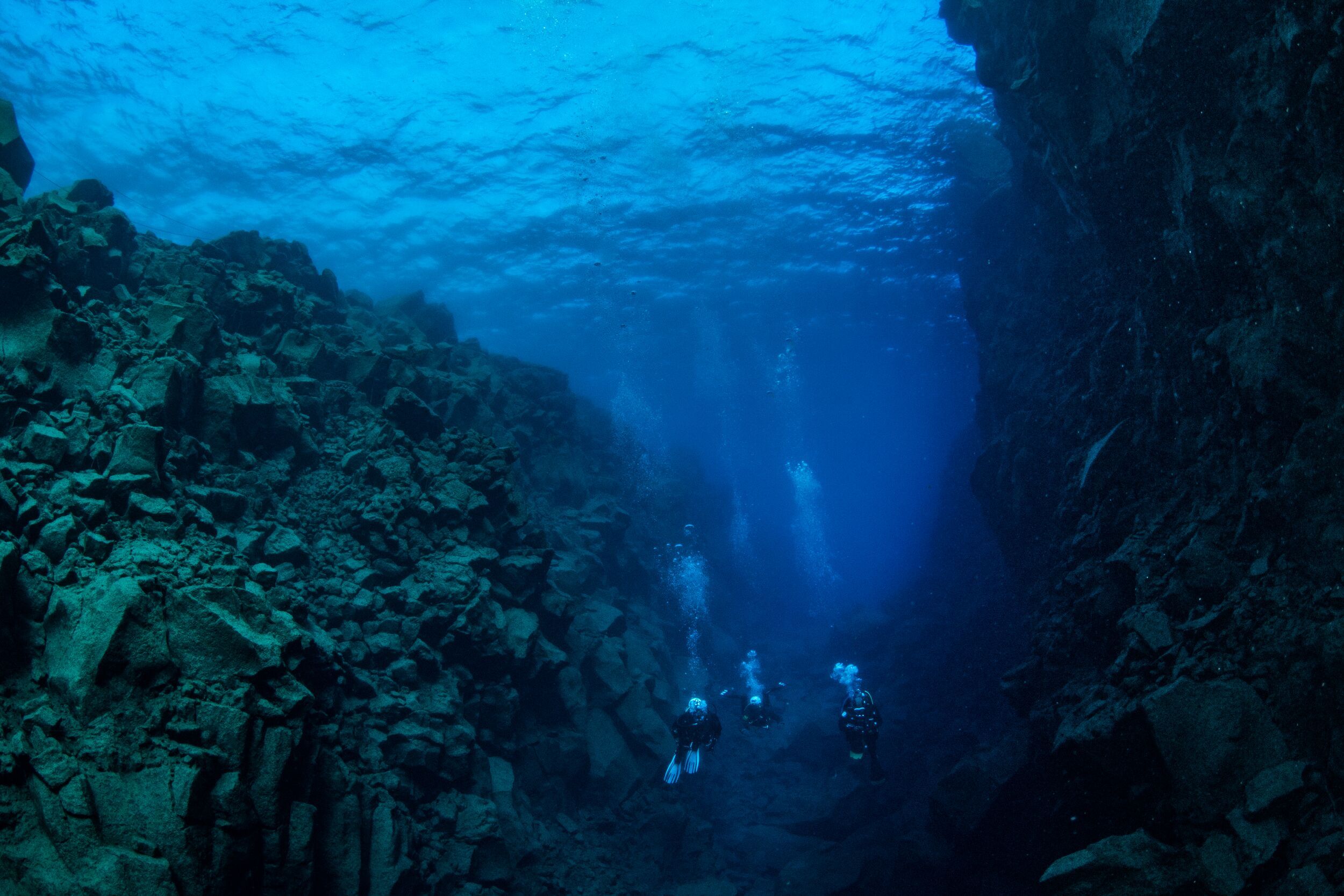 Divers exploring a deep, rocky underwater canyon with clear blue water.