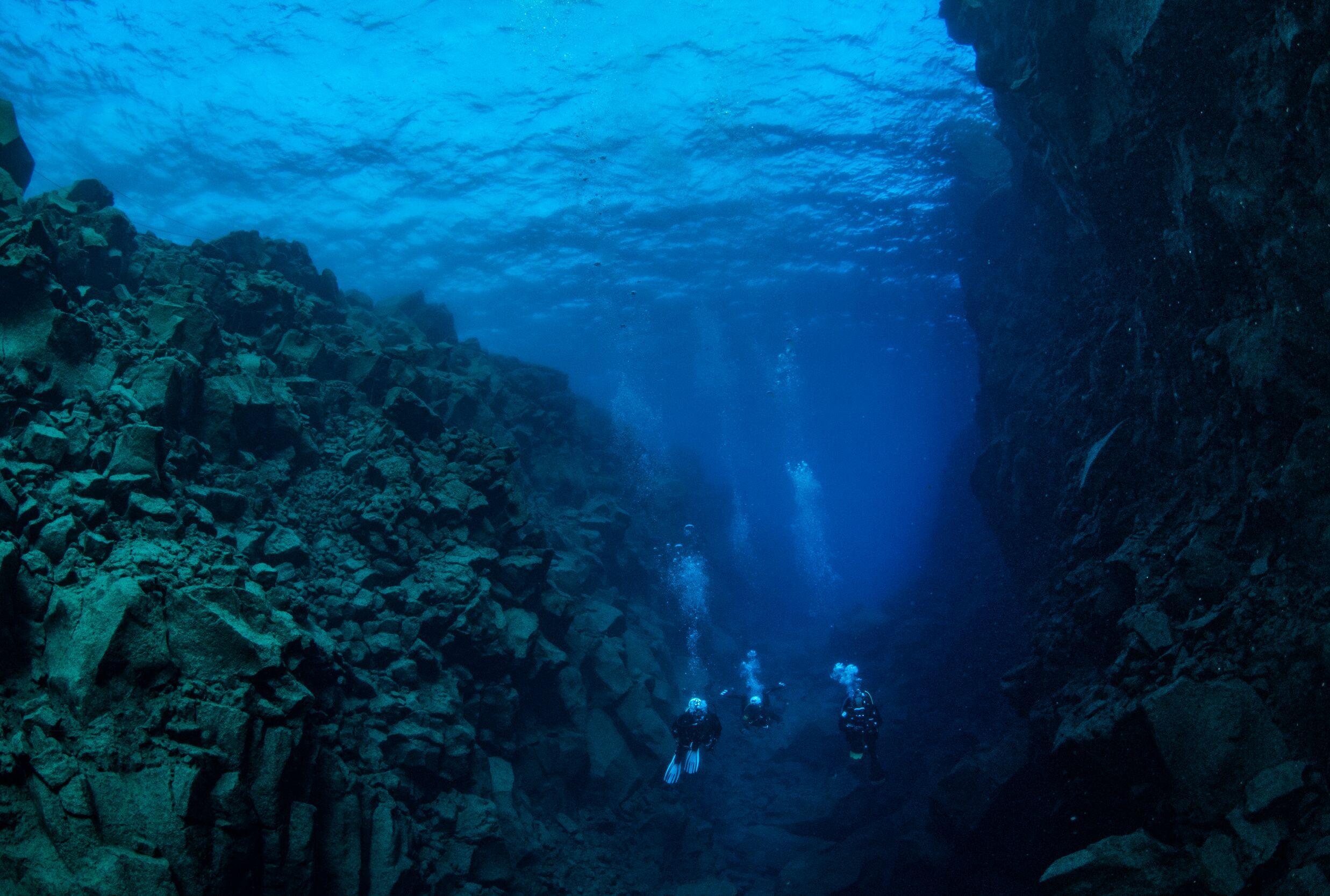 Divers exploring a deep, rocky underwater canyon with clear blue water.
