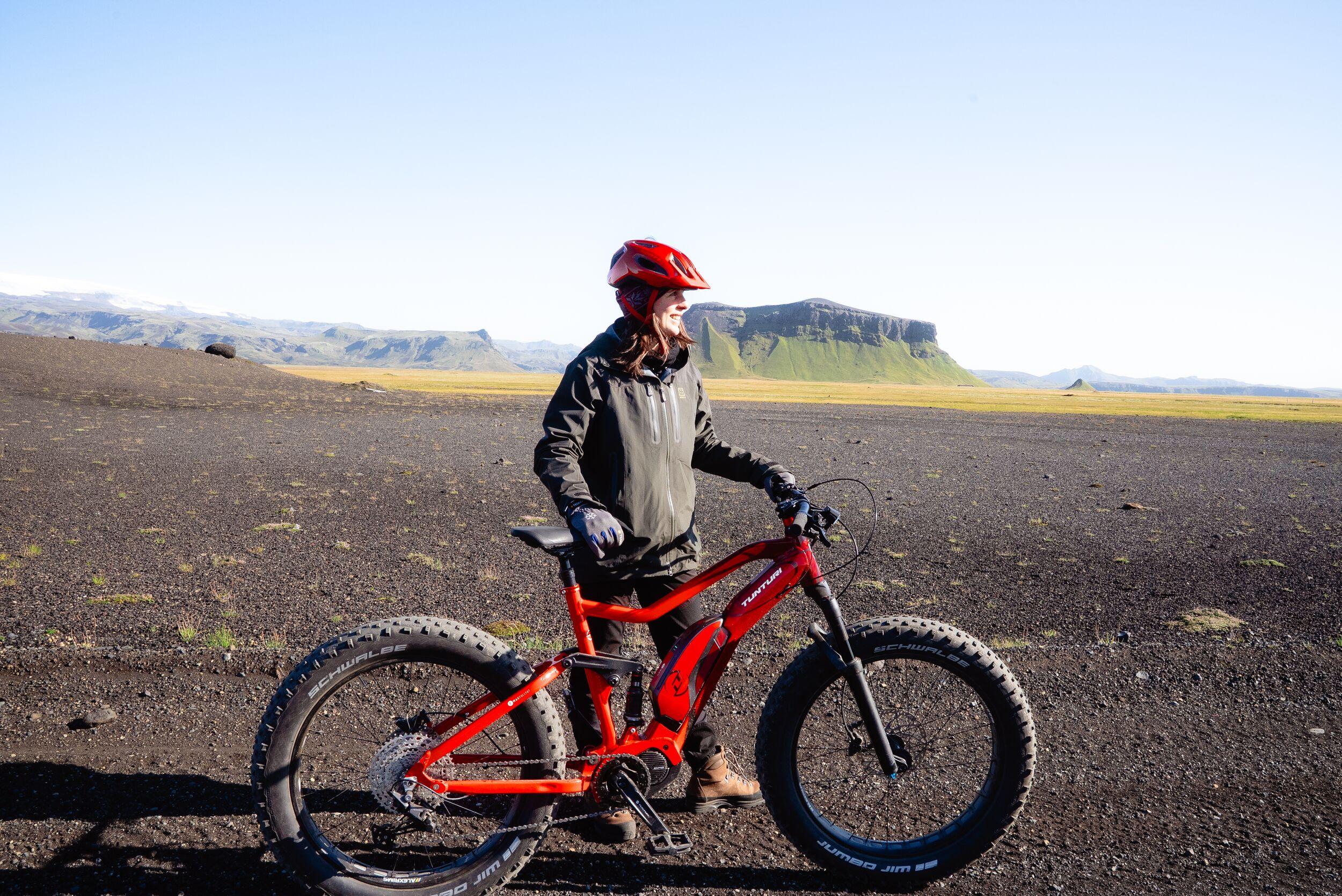 A person in a red helmet stands next to a red fat-tire electric bike on a dark, rocky plain with green-topped mountains in the background.