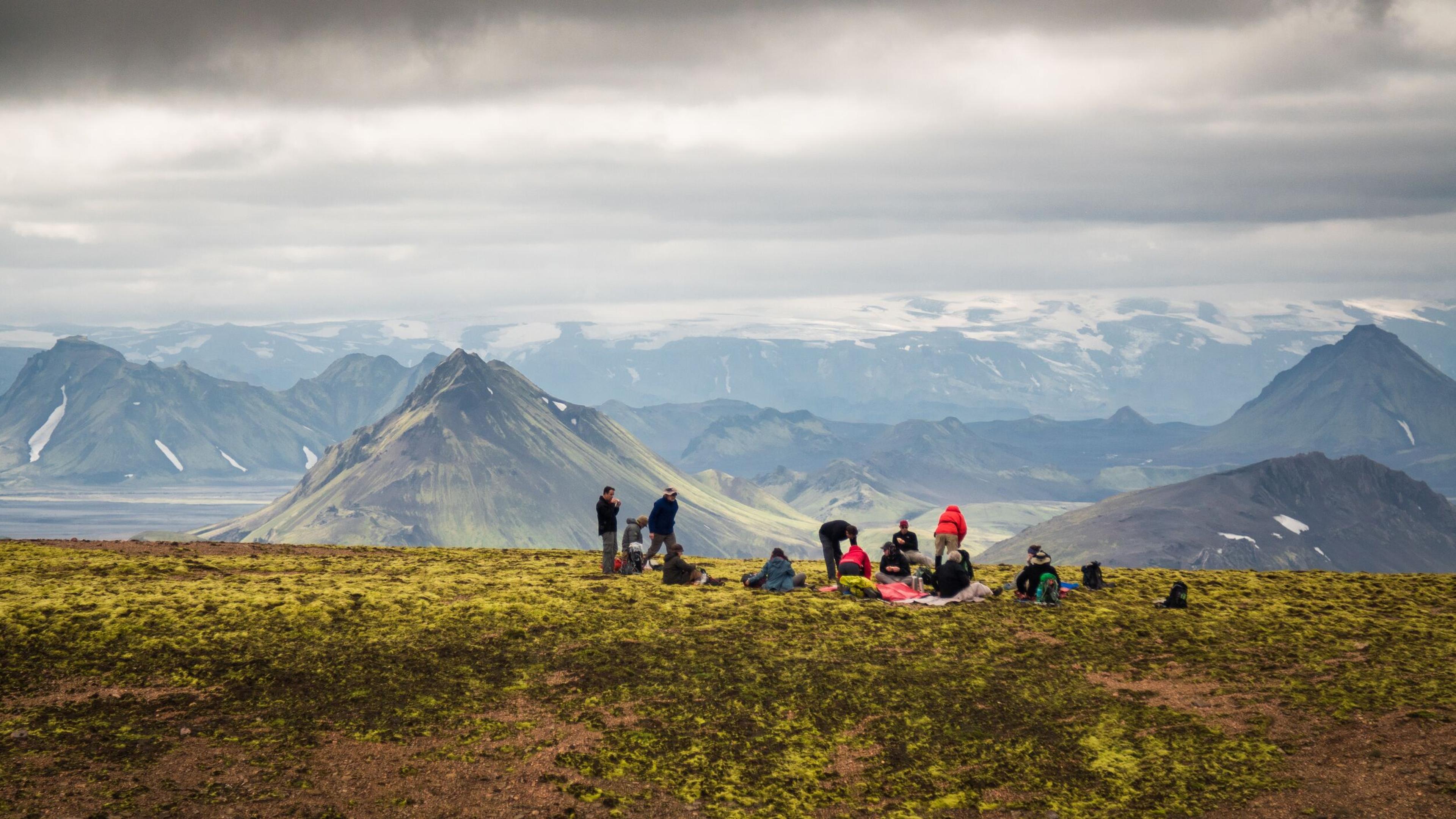 Hikers resting on a mossy slope overlooking a mountain range with distant glaciers under an overcast sky.