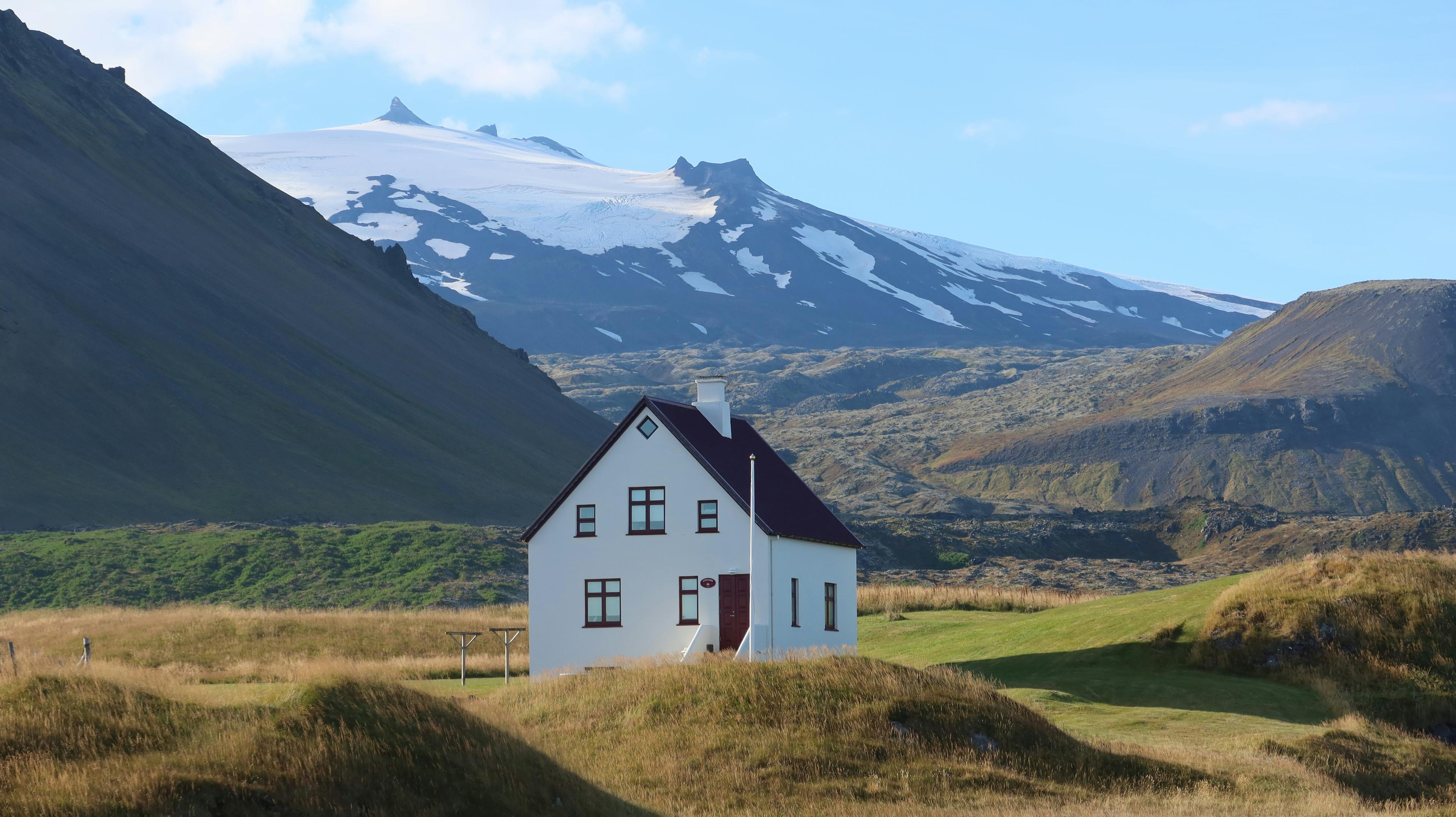 A charming white house with a red roof stands alone in a grassy Icelandic landscape, with the snow-capped Snæfellsjökull glacier and rugged mountains towering in the background.