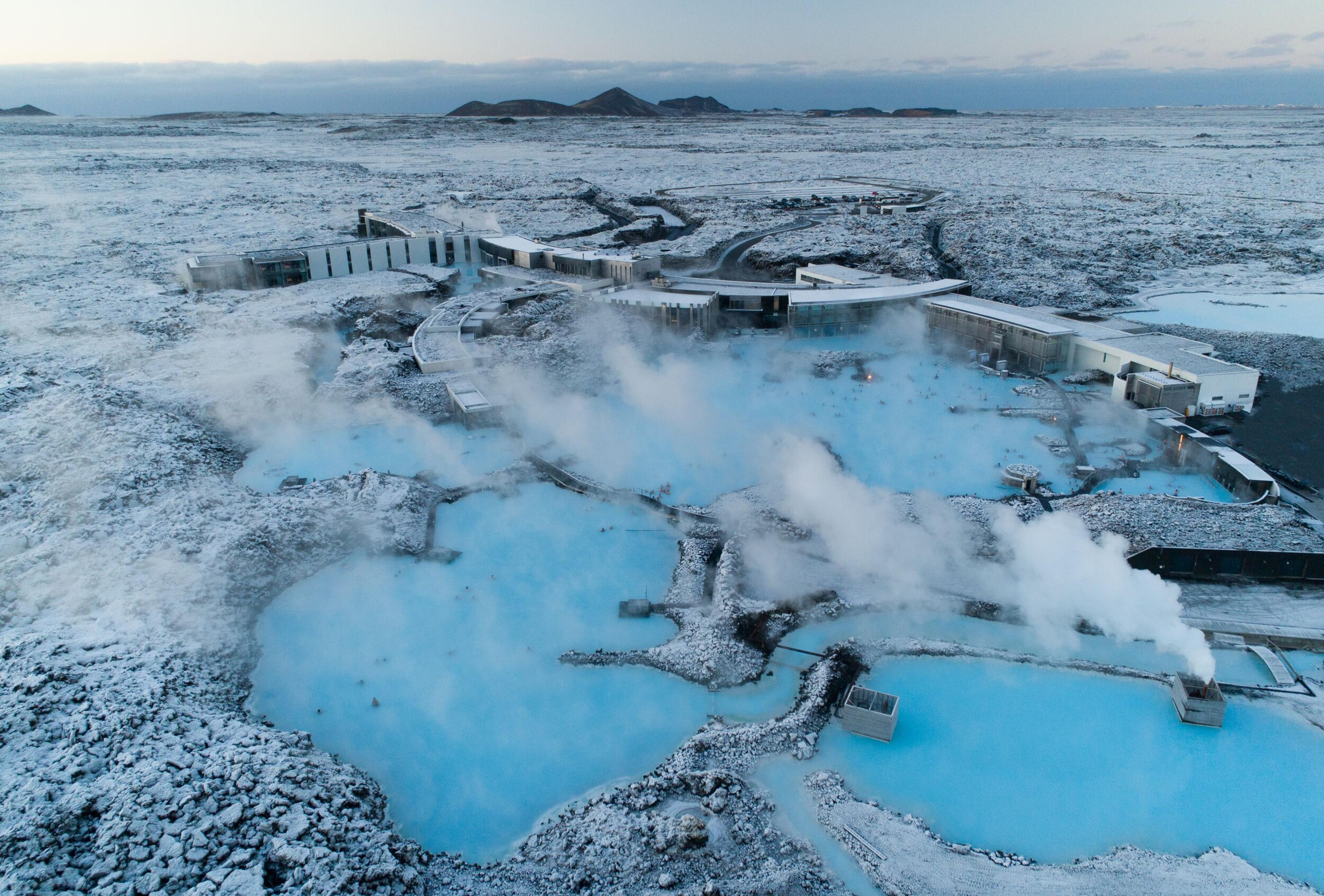 Aerial view of Iceland's Blue Lagoon, featuring steaming, bright blue geothermal pools surrounded by snow-covered volcanic rock and buildings.