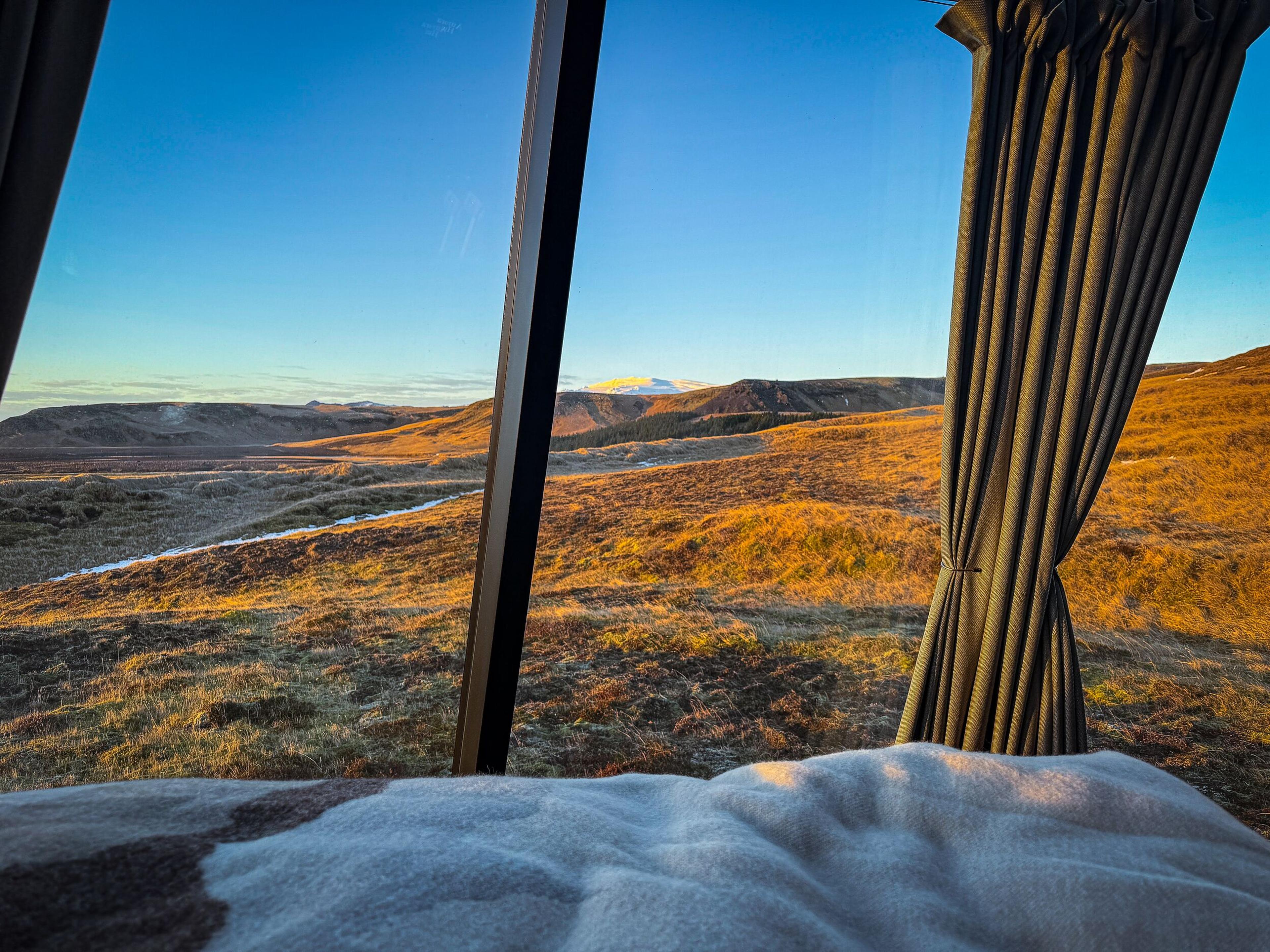 Sunrise view from a bed through a window showing a golden, hilly landscape with a stream and distant snowy peak.
