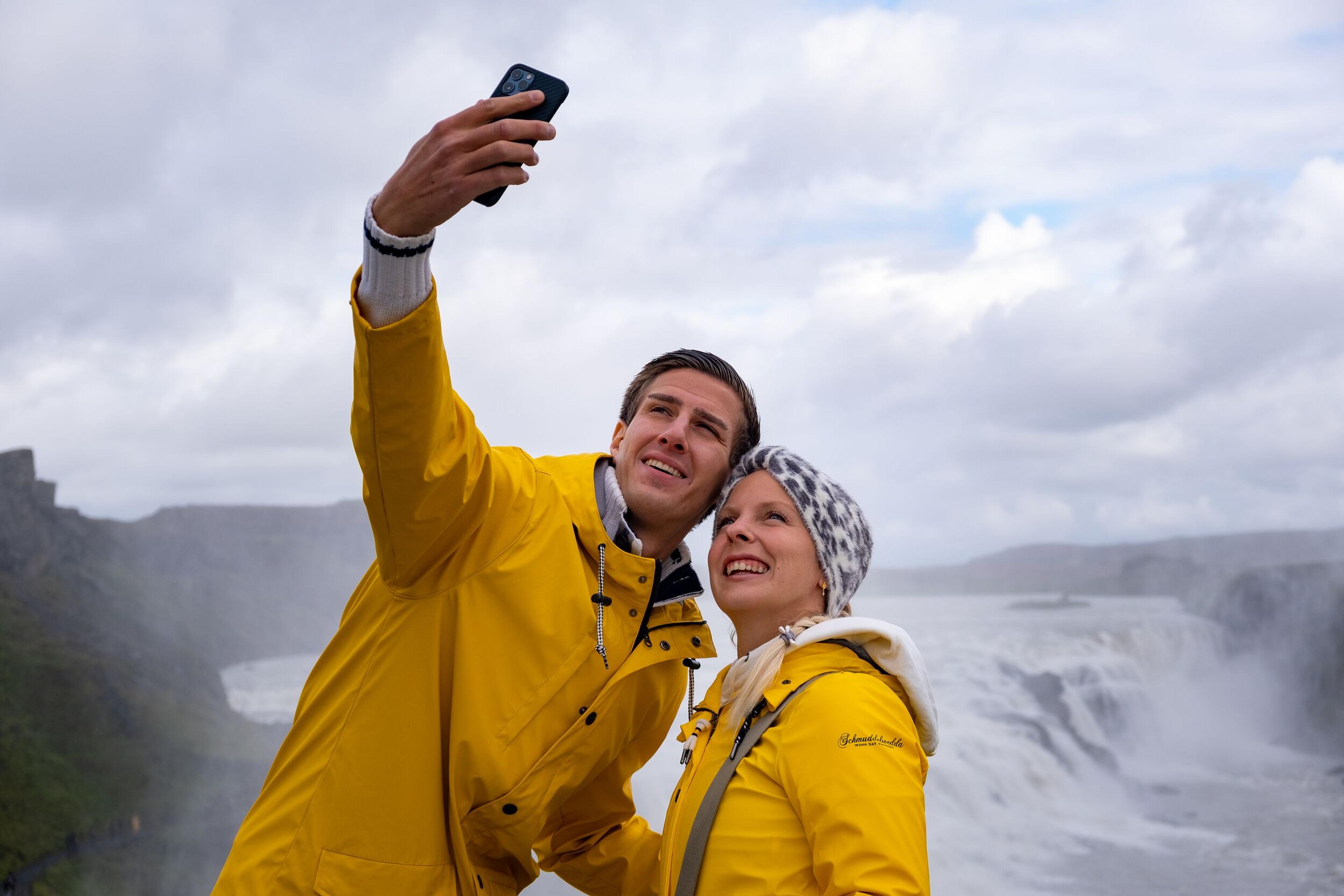 Two people in matching yellow raincoats are taking a selfie with a waterfall in the background, sharing a joyful moment in a dramatic natural setting.