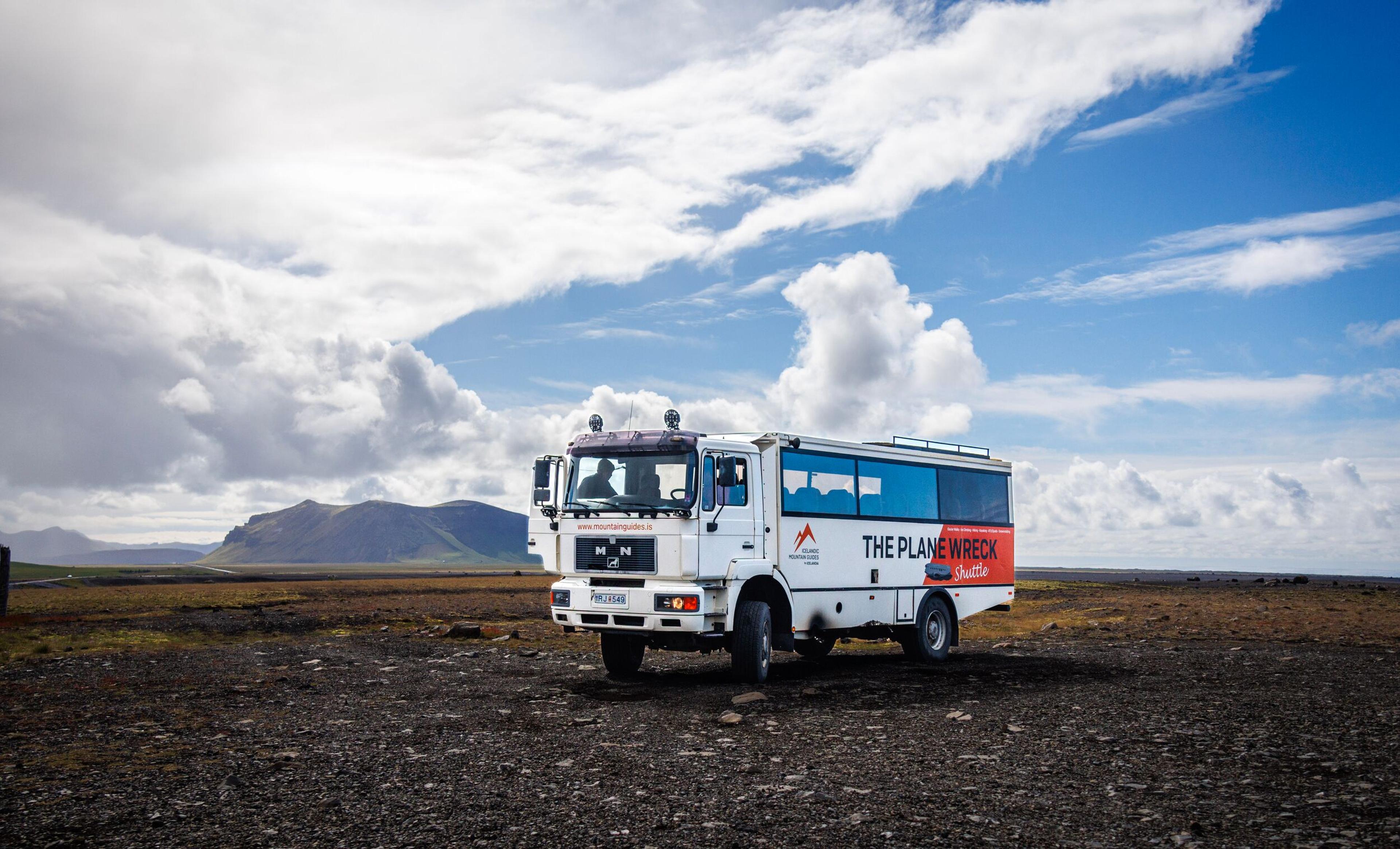 A white tour bus labeled "THE PLANE WRECK" on its side sits in a dark, rocky landscape with mountains under a partly cloudy sky.