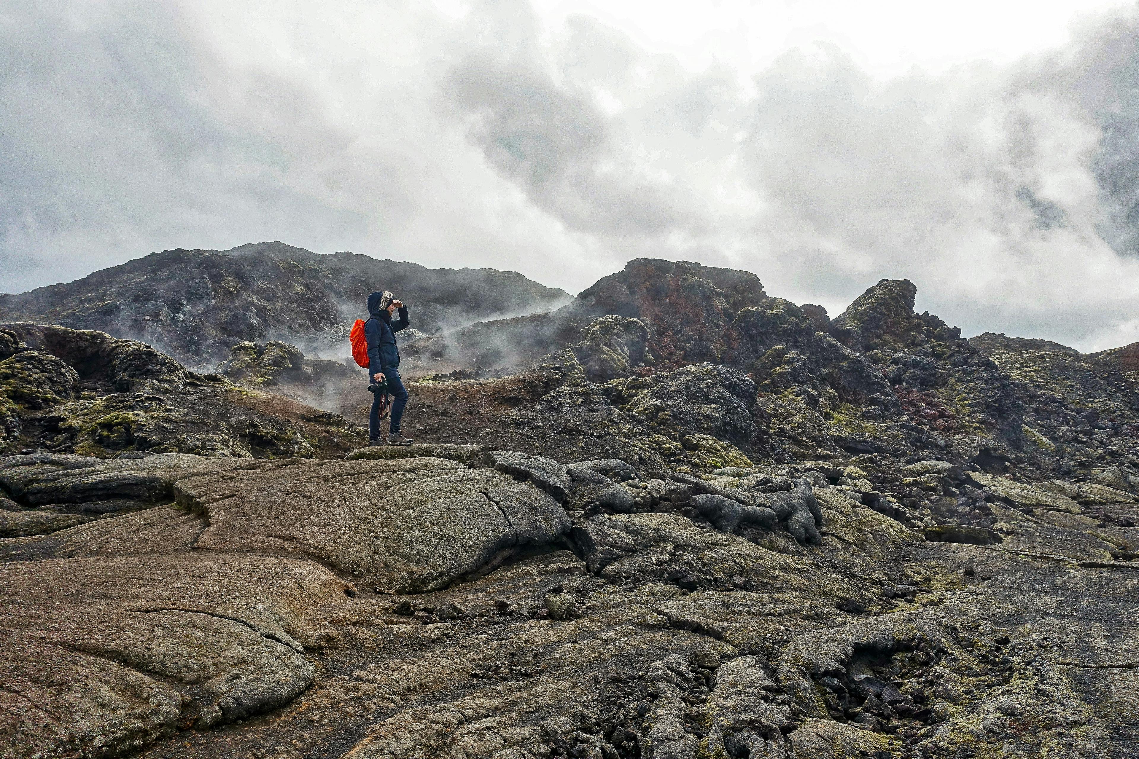 Person hiking near the steaming Krafla volcano, wearing dark clothing and a red backpack, surrounded by rugged volcanic terrain.