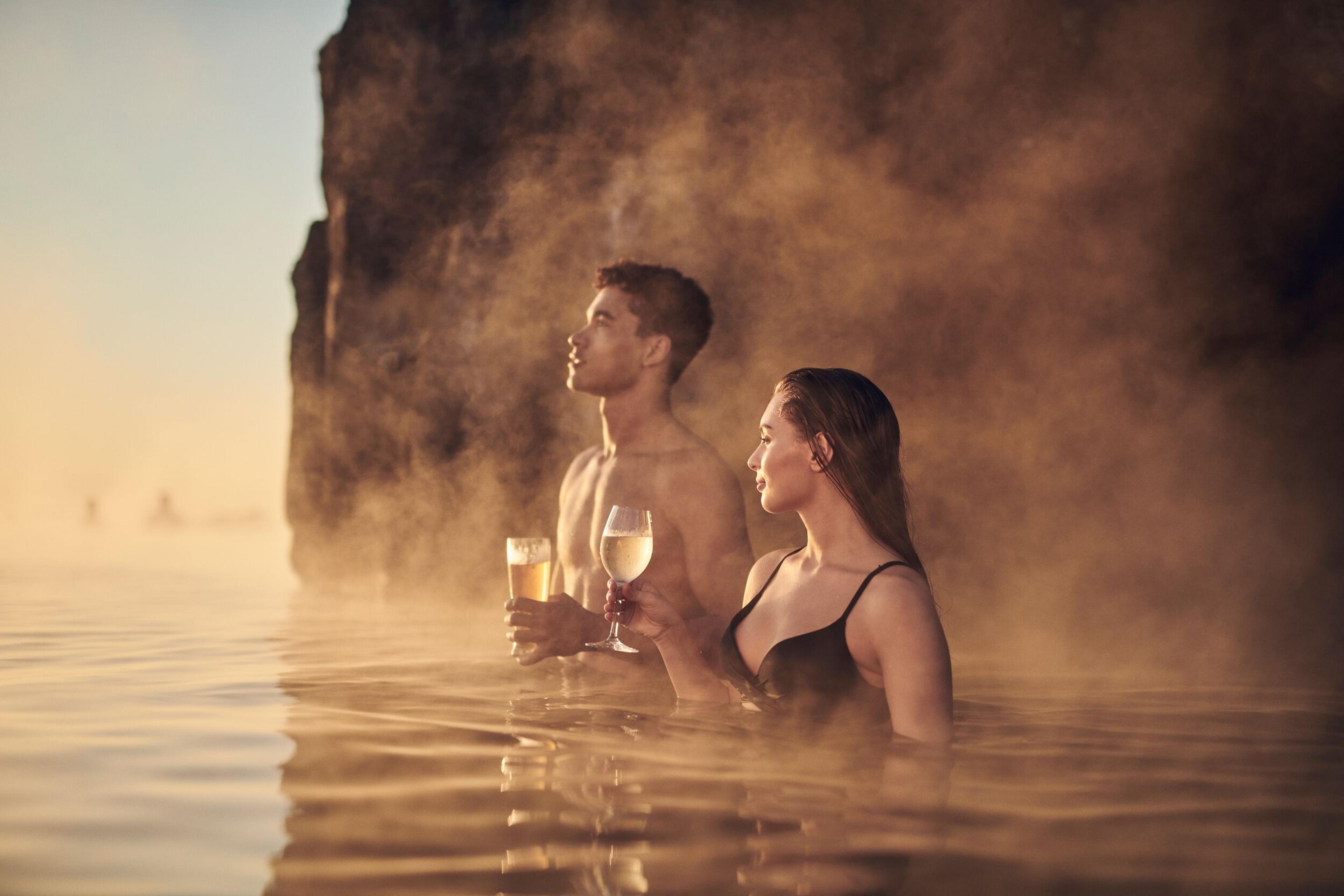 A couple with glasses of wine enjoying a serene moment in the warm, misty waters of Sky Lagoon, with a glowing sunset light illuminating their faces and the rocky cliff behind them.