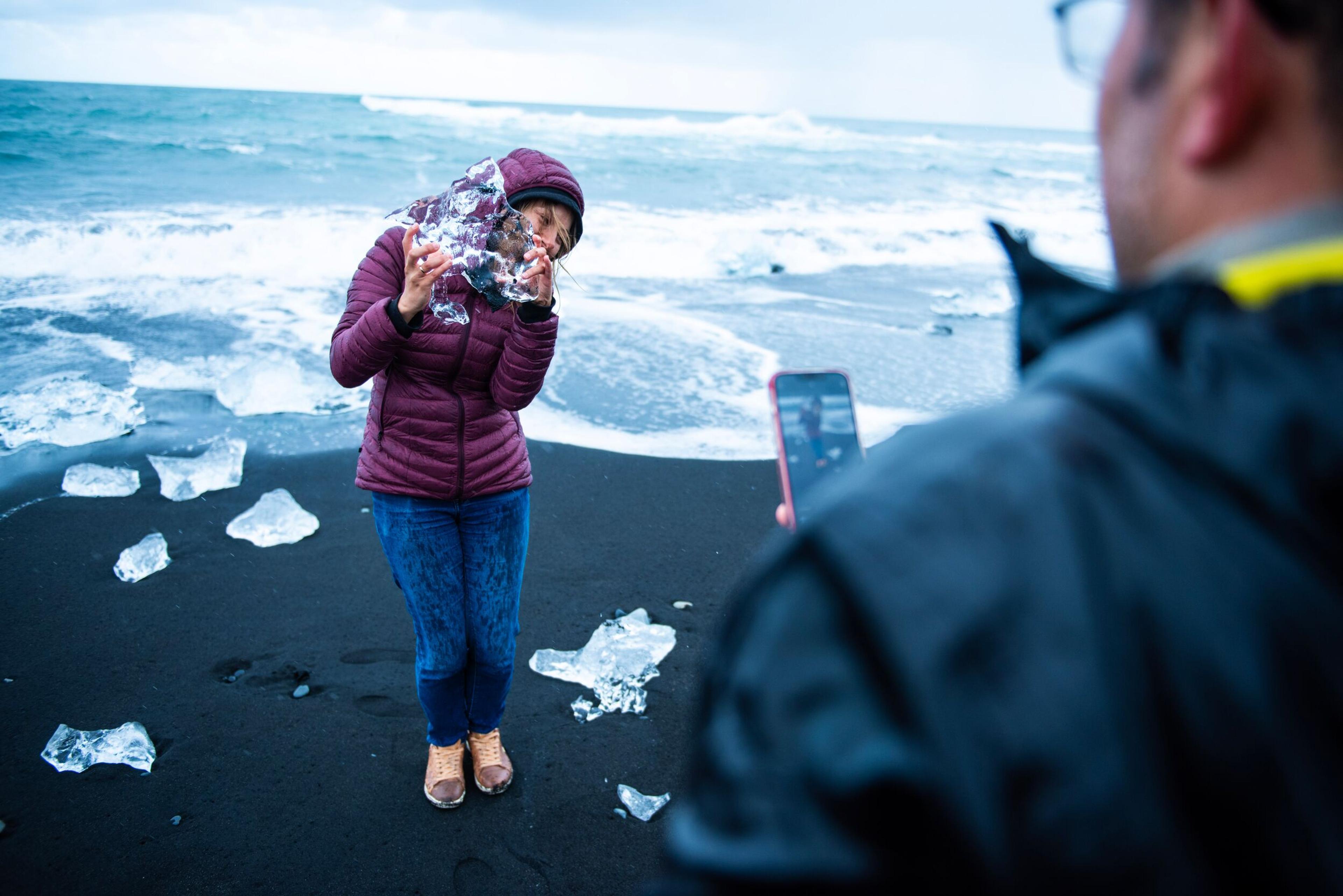 A woman poses with a large chunk of ice on a black sand beach, as a man photographs her with a phone.