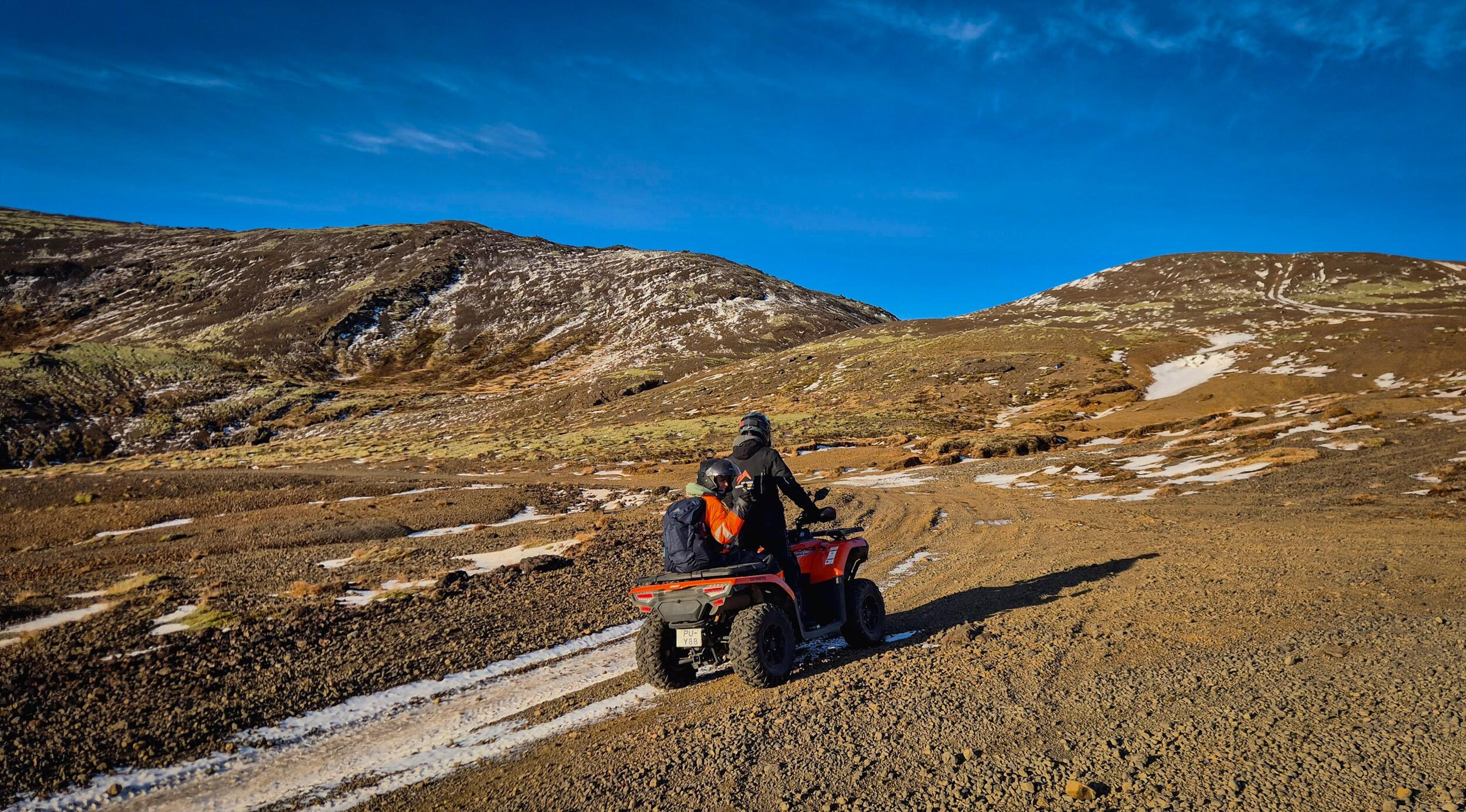 Two people ride an orange ATV on a dirt path through a rocky, snow-dusted landscape.
