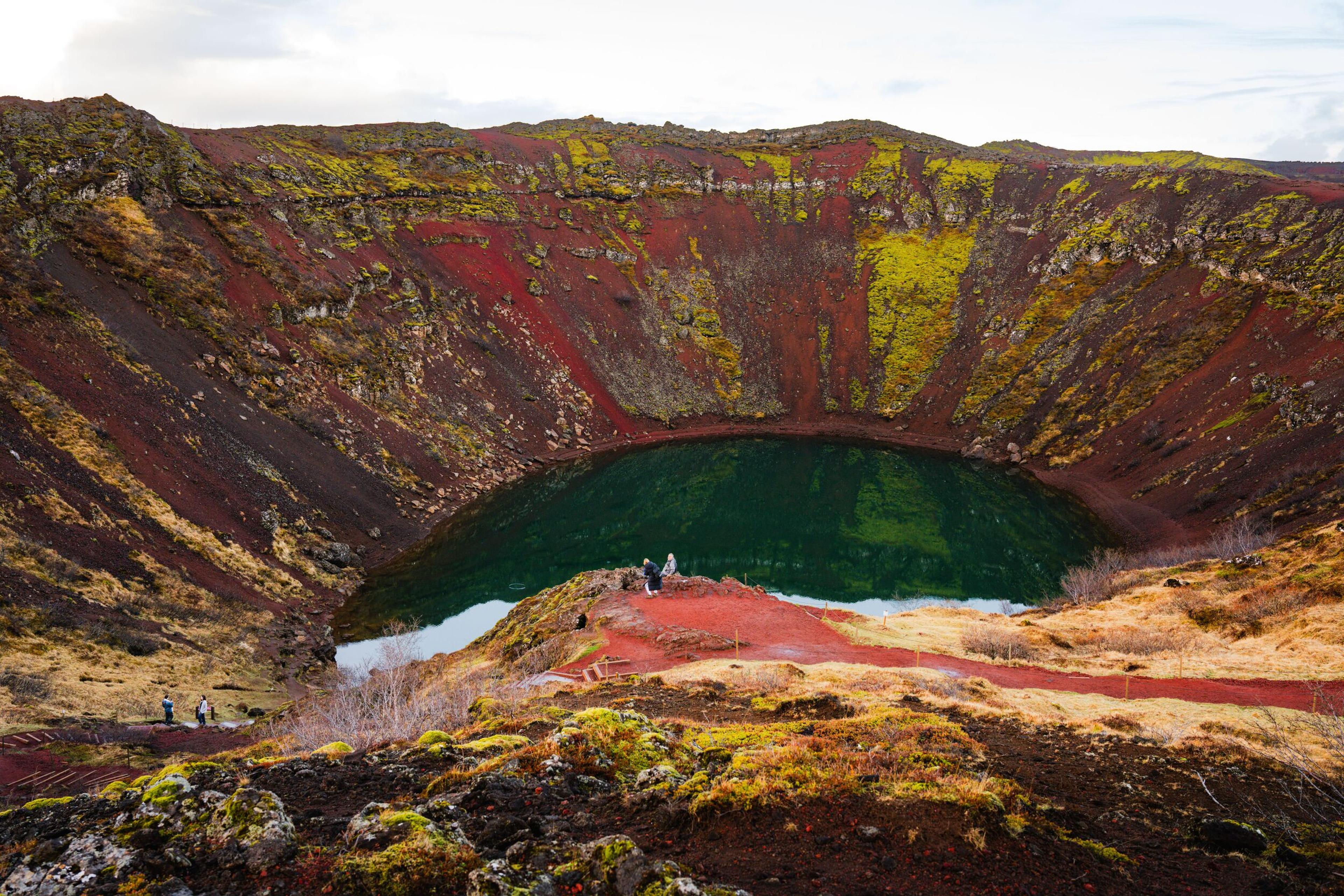 A volcanic crater with vibrant red and green moss-covered walls surrounding a striking blue-green lake, with a few people on the rim.