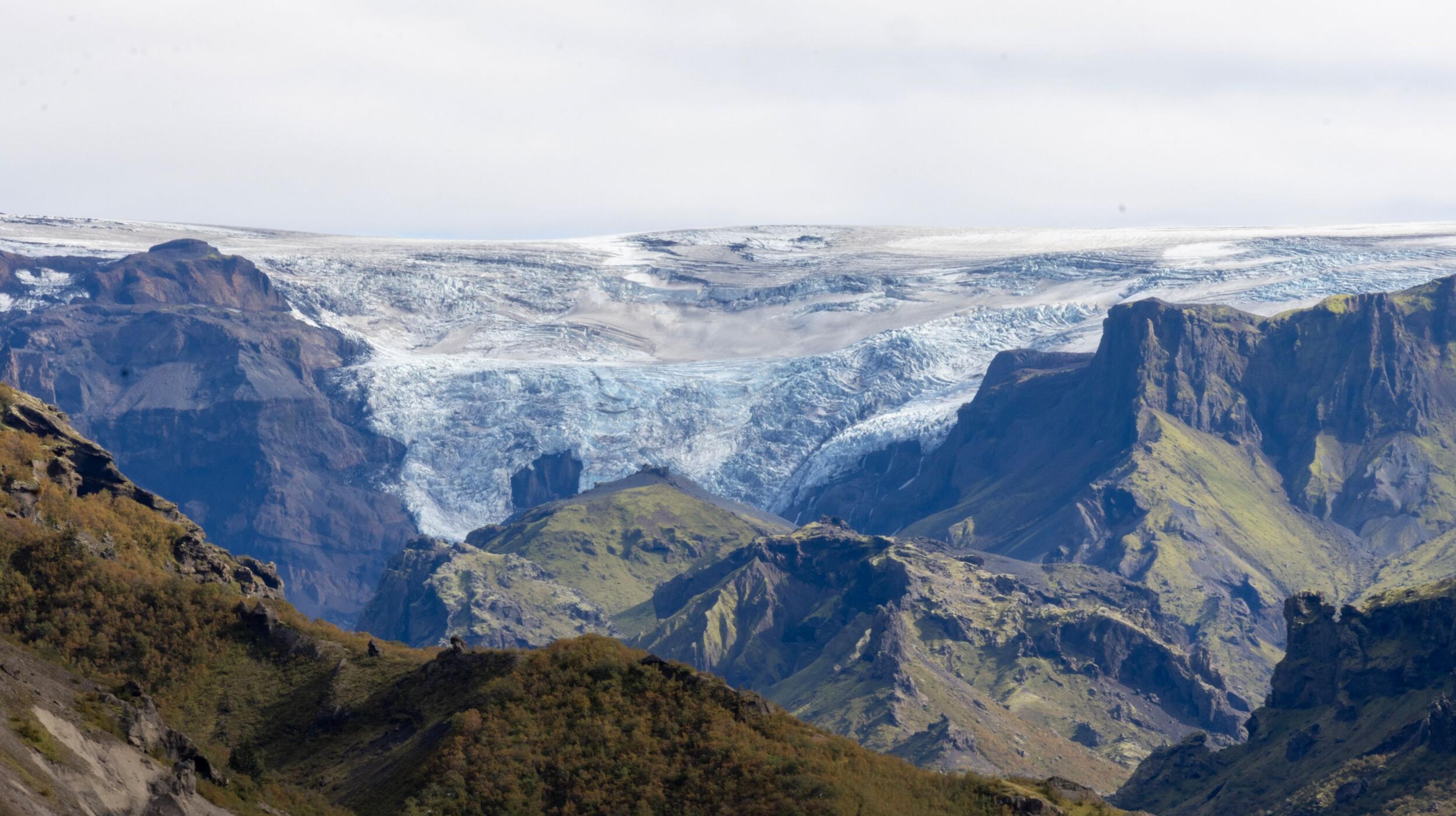 A vast glacier between rugged, vegetated mountains under a pale sky.