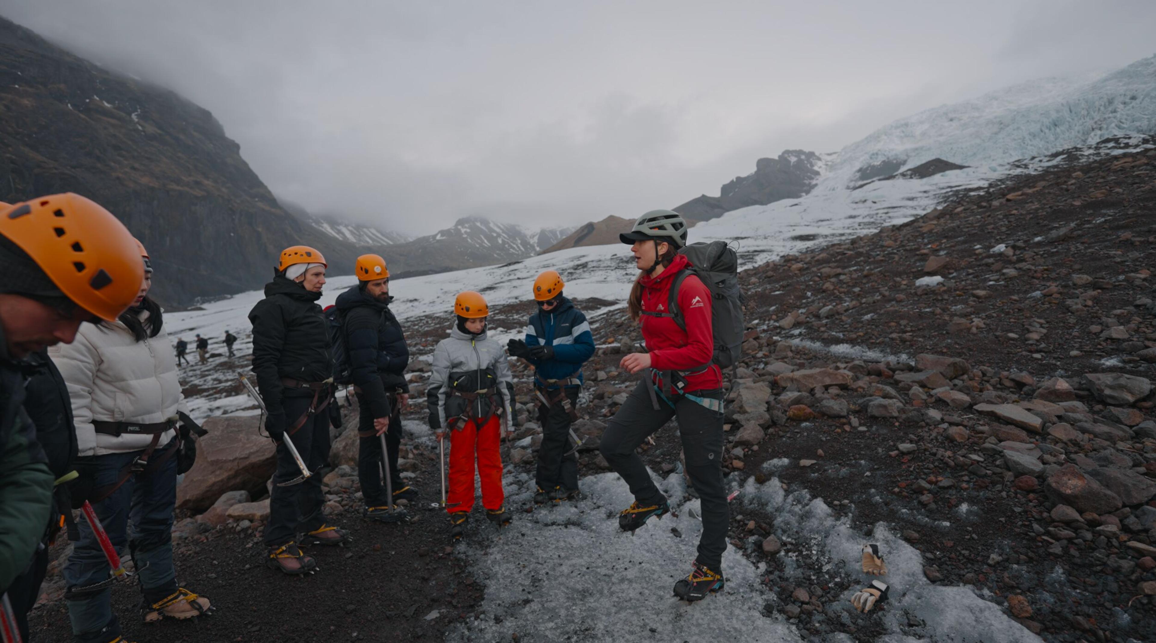 A guide in a red jacket addresses a group of glacier hikers on an icy, rocky terrain.