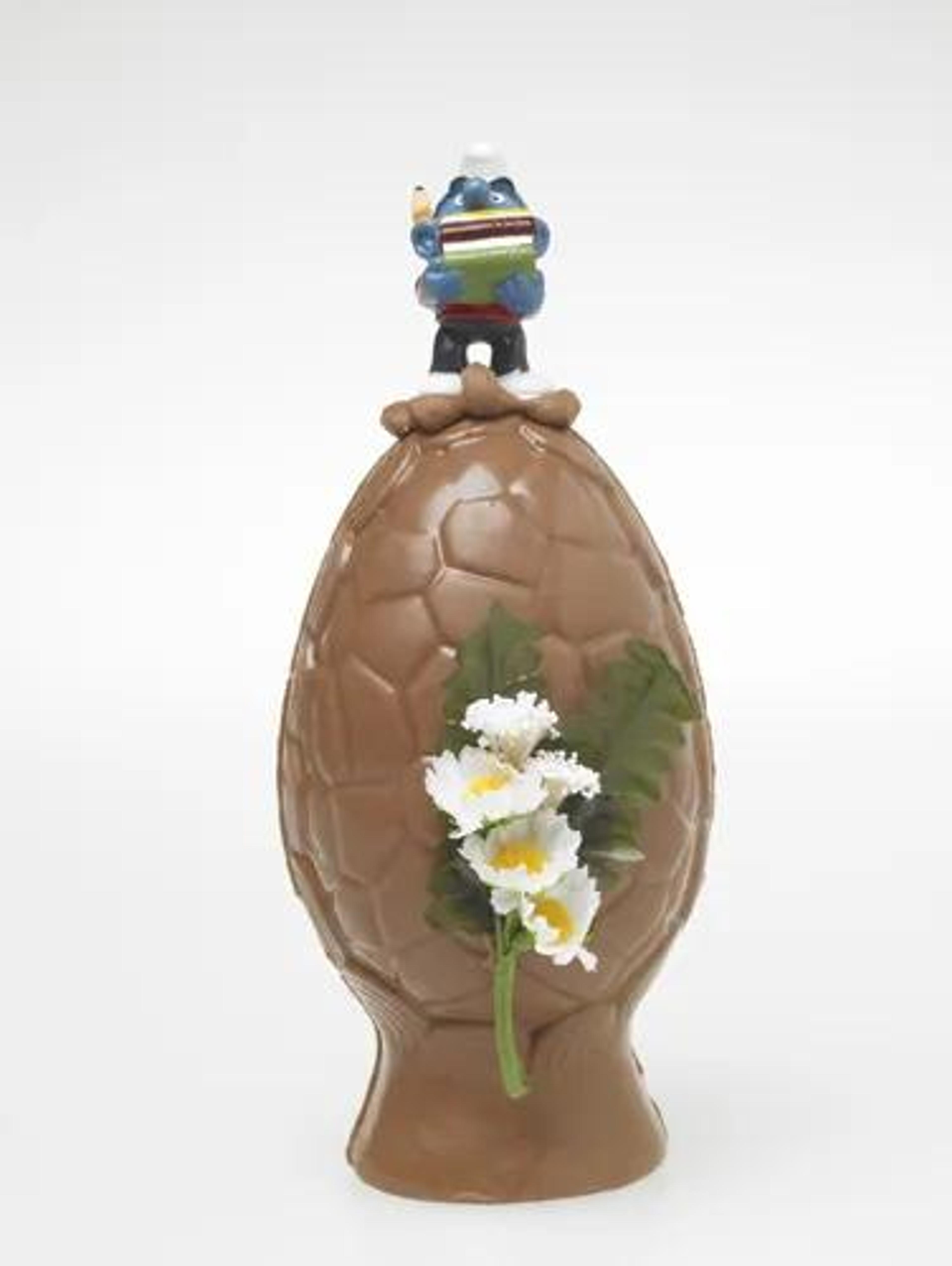 A chocolate Easter egg decorated with a blue chef figurine holding books on top and white flowers on the side.
