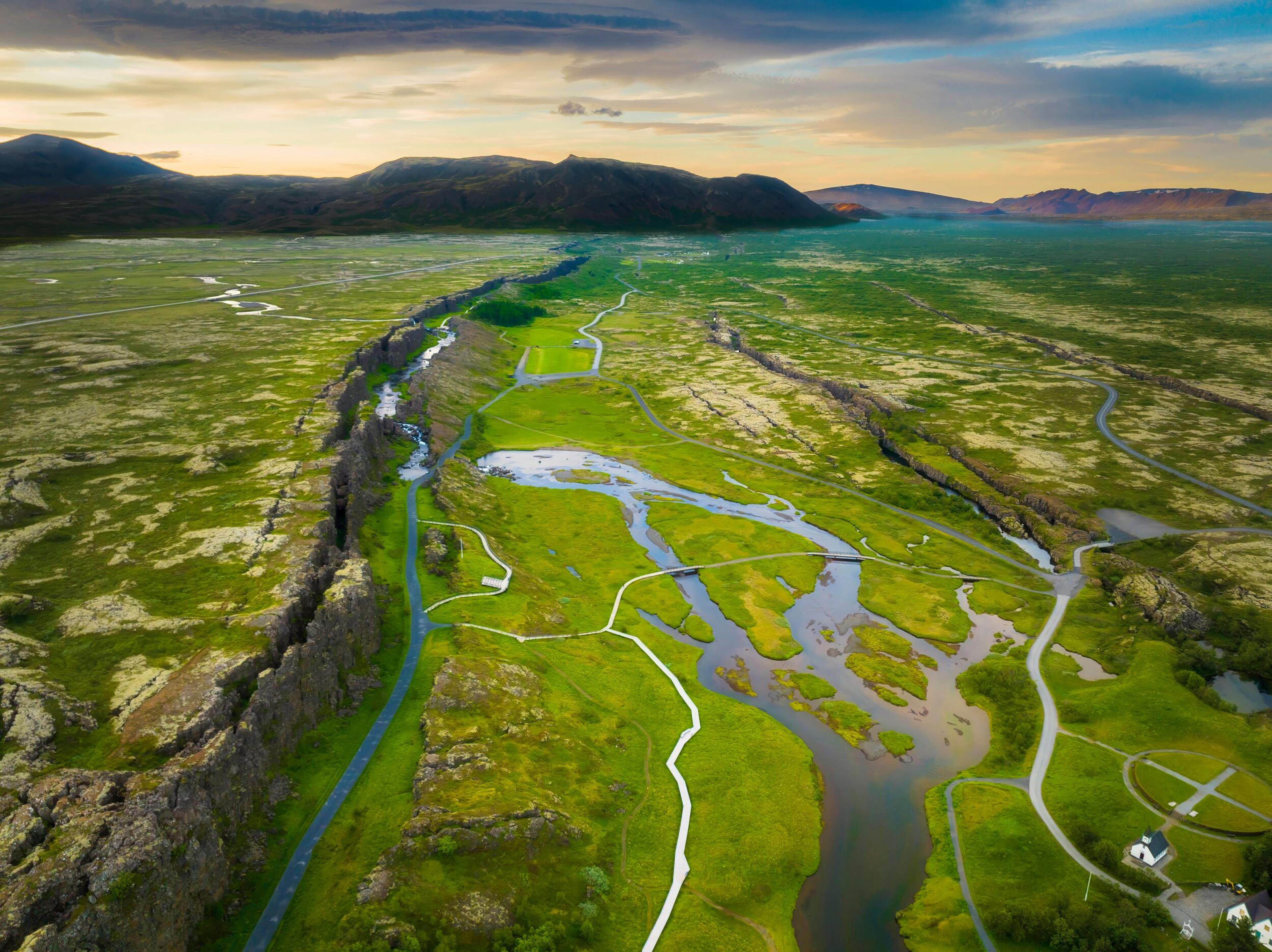 Aerial view of the lush, meandering waterways of Þingvellir National Park, flanked by the stark Almannagjá rift, under a dramatic Icelandic sky.