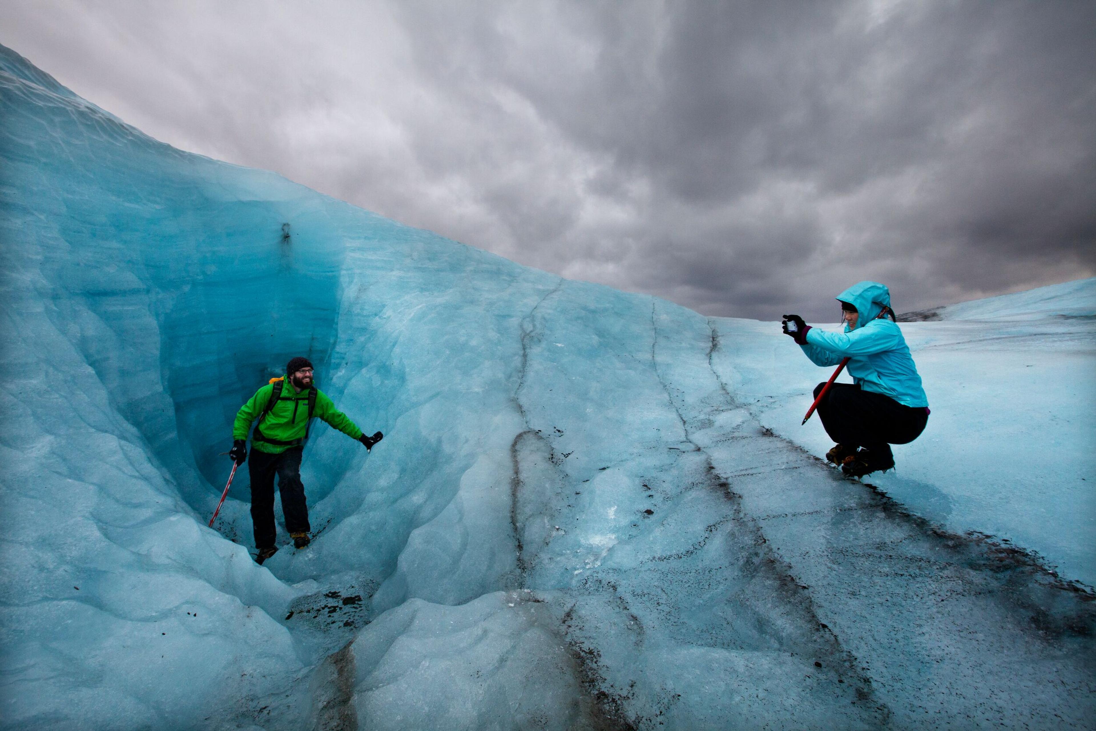 Two individuals on a blue glacier; one capturing a photo while the other strikes a pose.