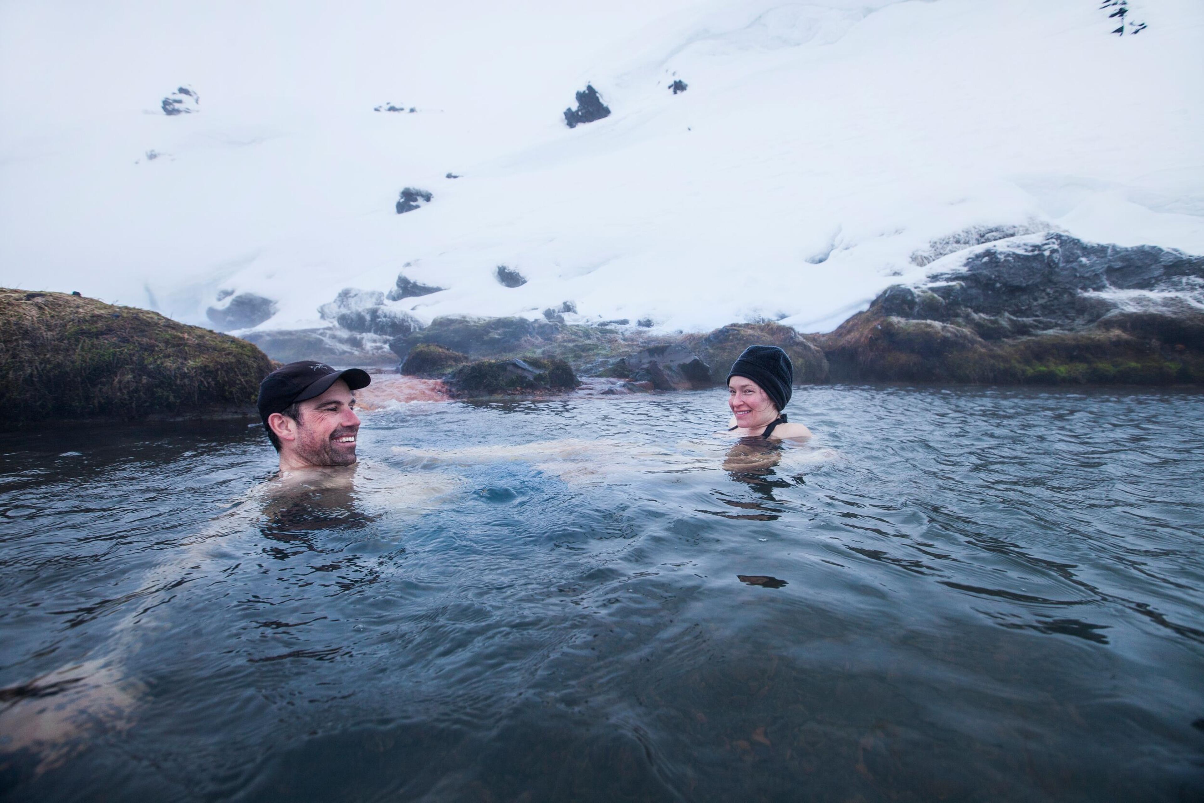 Two people in winter caps relaxing in the hot spring at Landmannalaugar surrounded by snowy rocks.