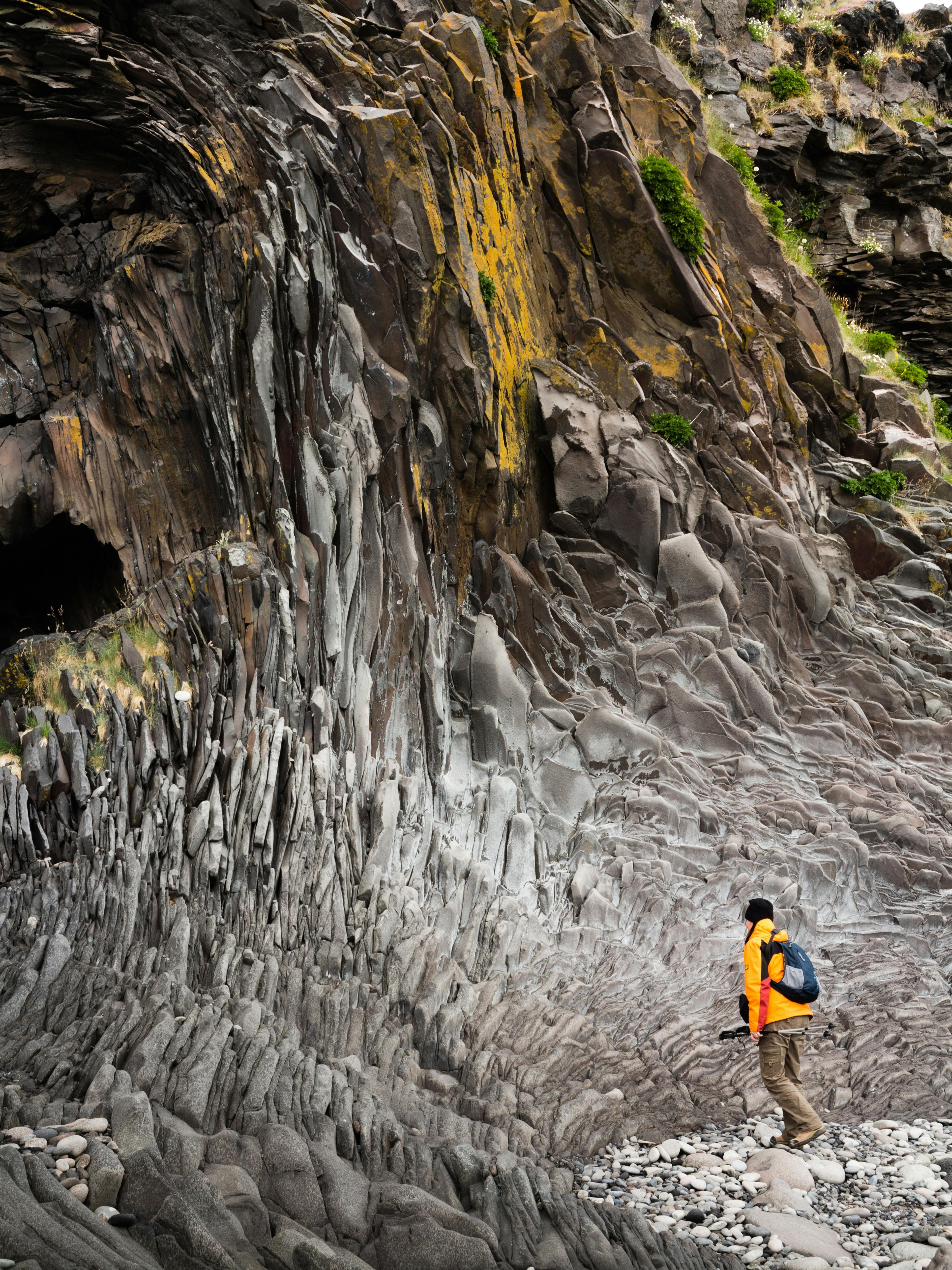 A person in a bright orange jacket explores a striking geological formation with sharp, jagged rocks and layers of smooth, wave-like stone patterns, set against a backdrop of lush greenery and moss.