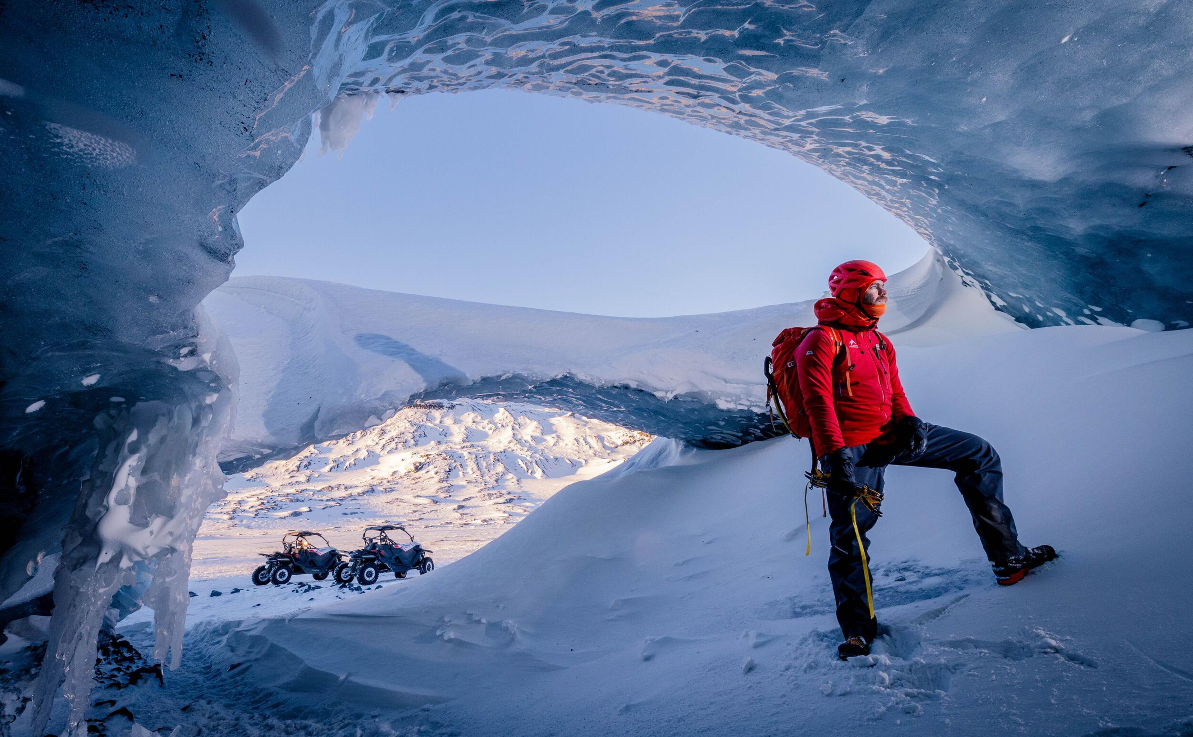 A person dressed in red winter gear stands inside the Askur Ice Cave, framed by ice formations with a clear view of snow-covered mountains and buggies in the background.