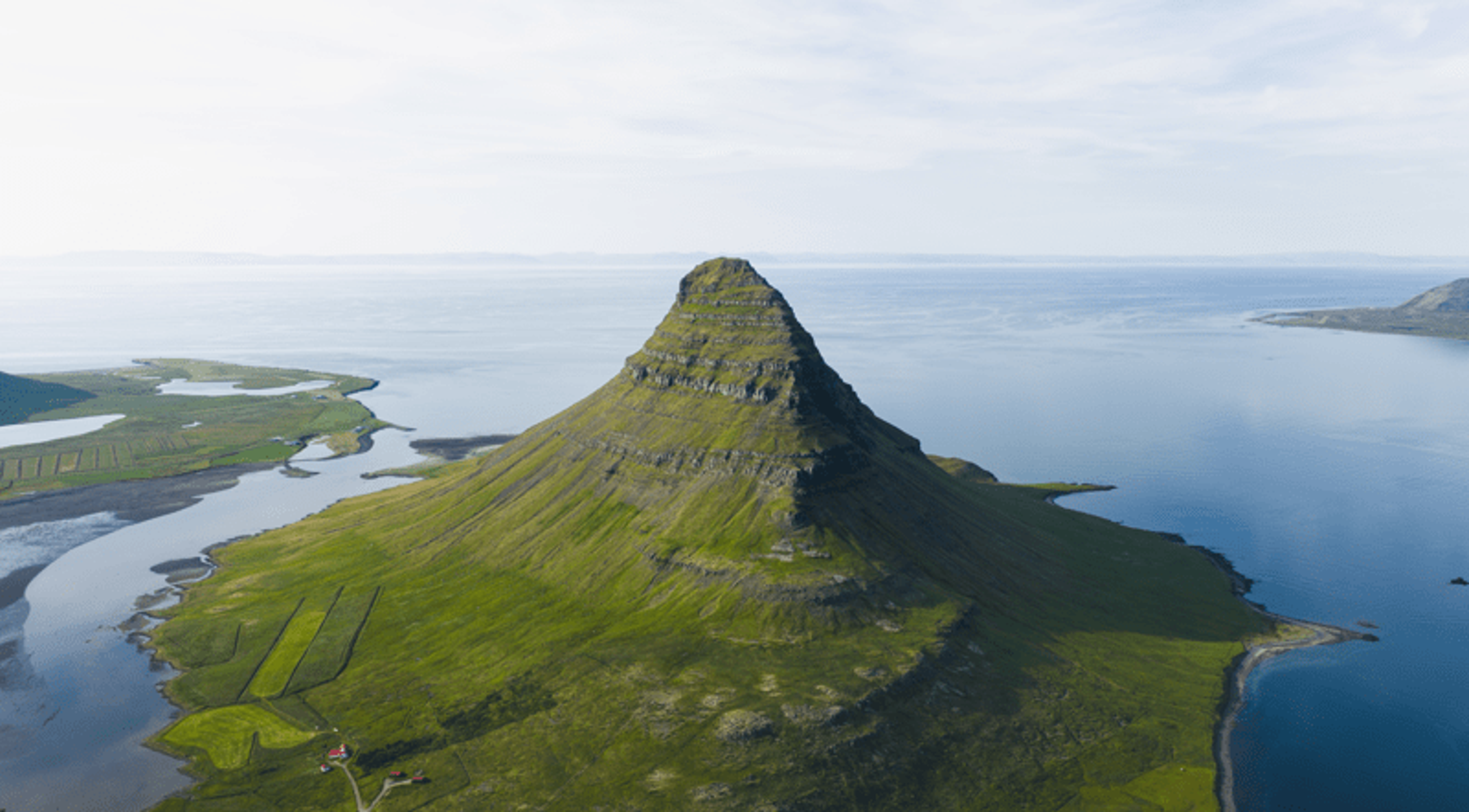 Aerial view of Kirkjufell, a conical green mountain, surrounded by water.