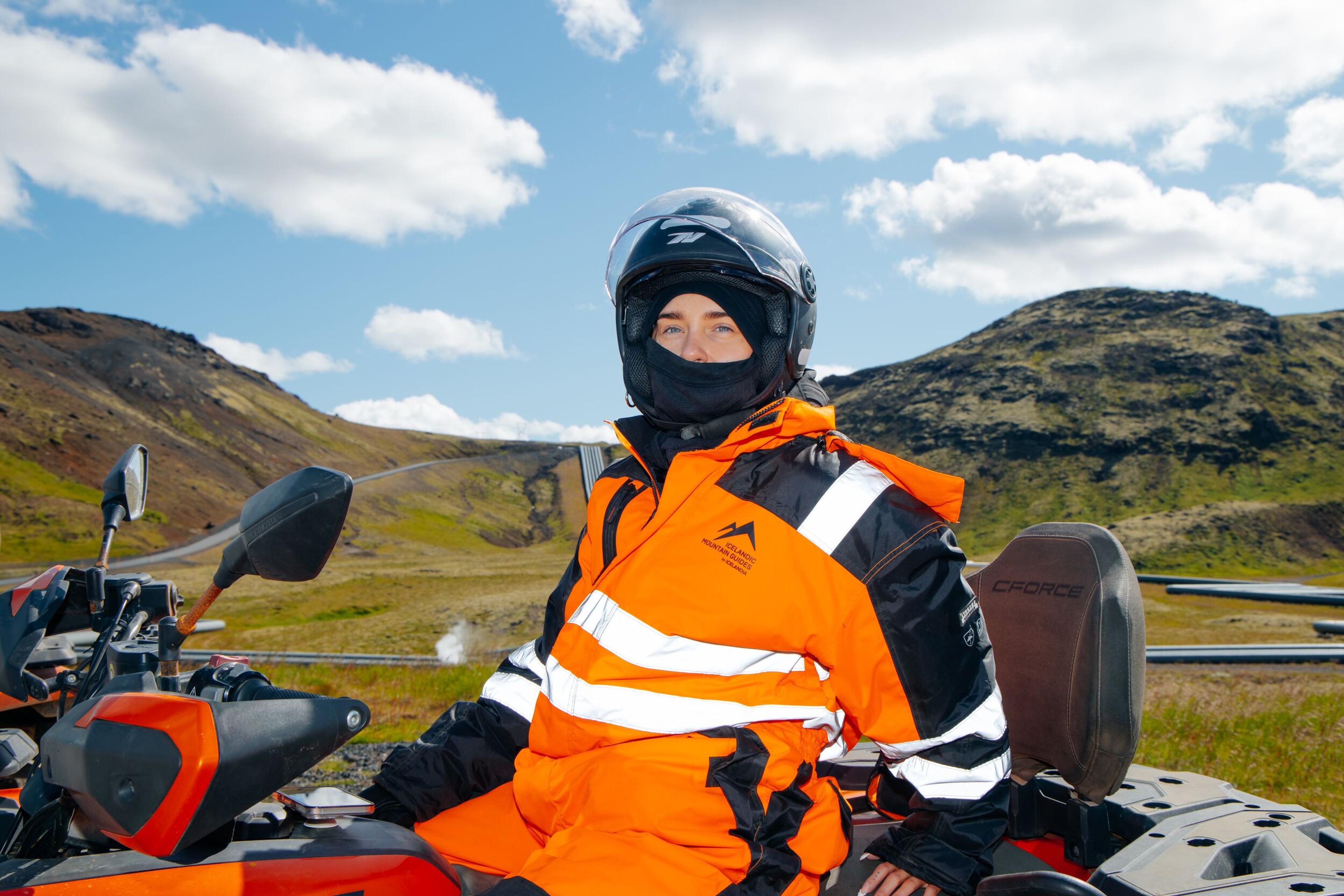 A person in an orange ATV suit and helmet sits on an ATV in a rugged, hilly landscape.